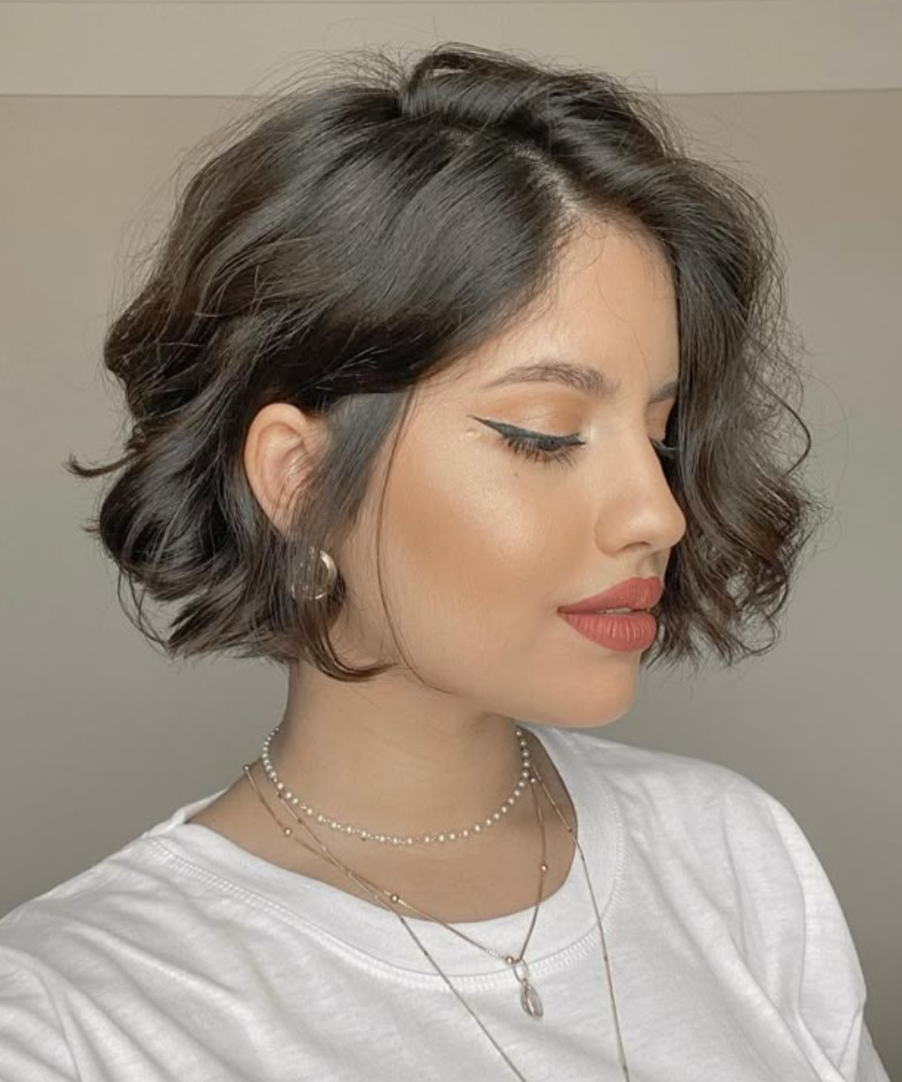 Corte de cabelo ondulado e curto destaca-se em perfil com maquiagem elegante e acessórios discretos.