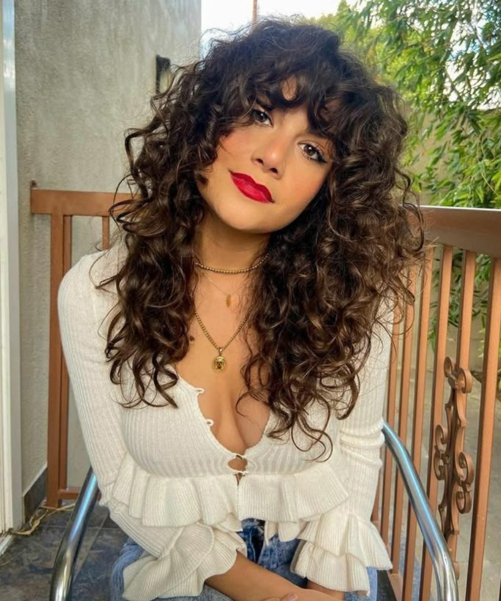 Mulher com corte de cabelo ondulado shaggy hair, usando batom vermelho, sentada em varanda com vegetação ao fundo.