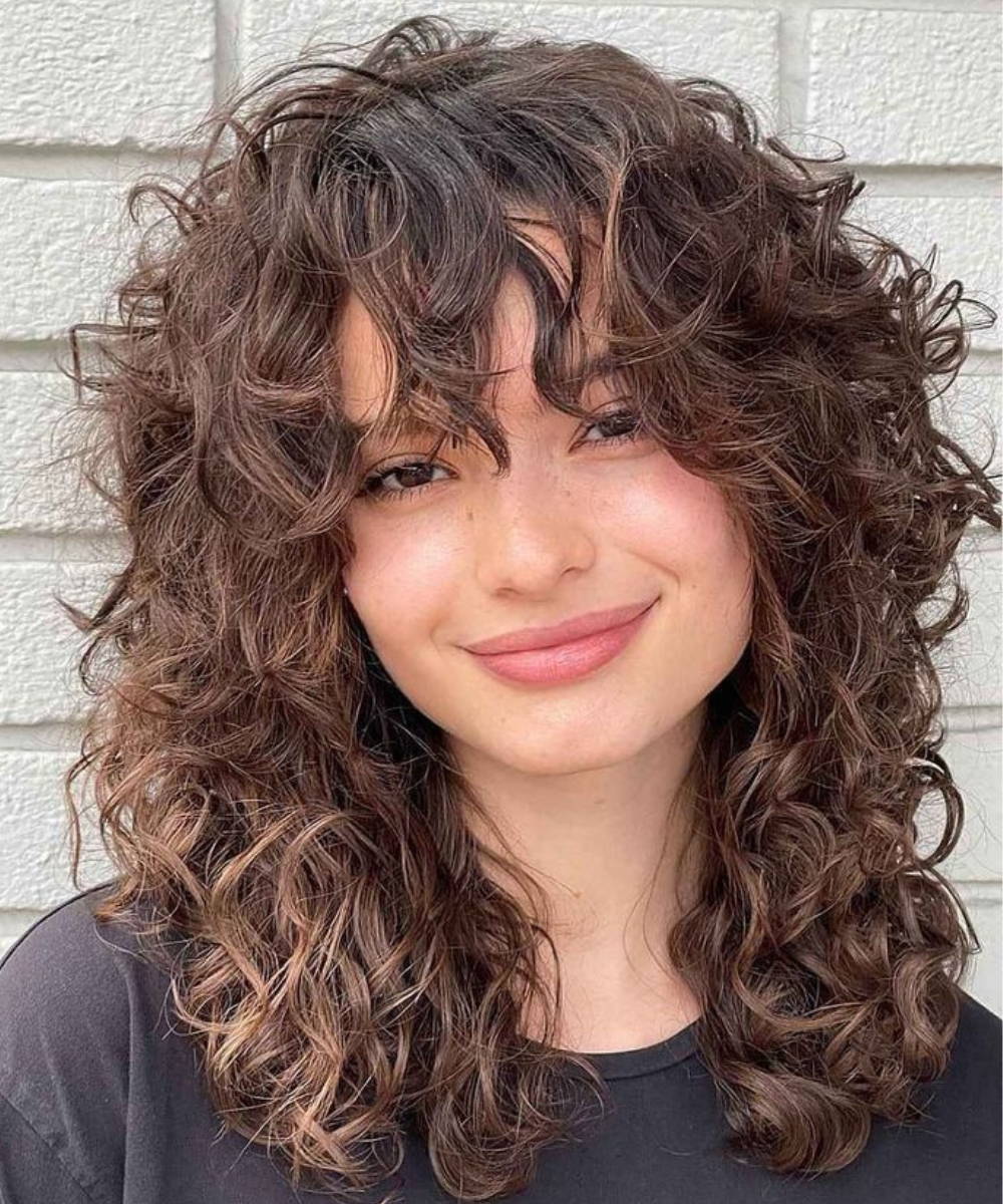 Mulher com corte de cabelo ondulado wolf cut, castanho e volumoso, sorrindo.