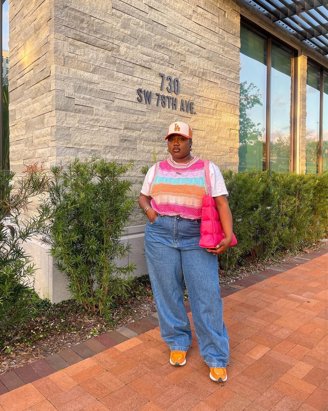 Pessoa com colete de crochê colorido e jeans soltos, segura bolsa rosa em rua de tijolos, ao lado de parede clara.