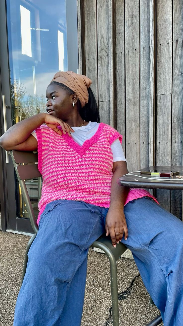 Pessoa sentada ao ar livre usando um colete de crochê rosa sobre uma camiseta branca, olhando para o lado.