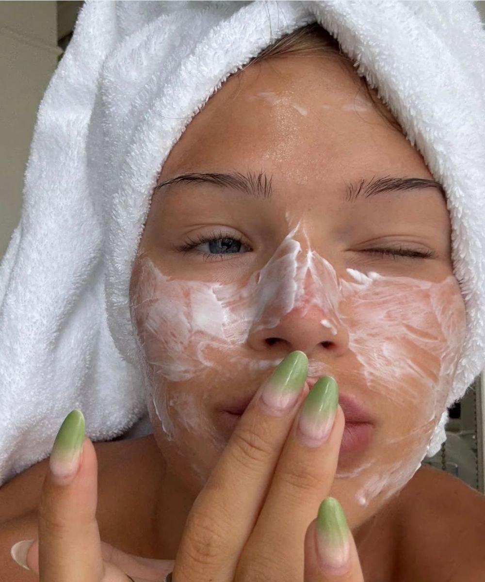 Pessoa com toalha na cabeça aplicando máscara facial, piscando um olho, com unhas compridas verdes.