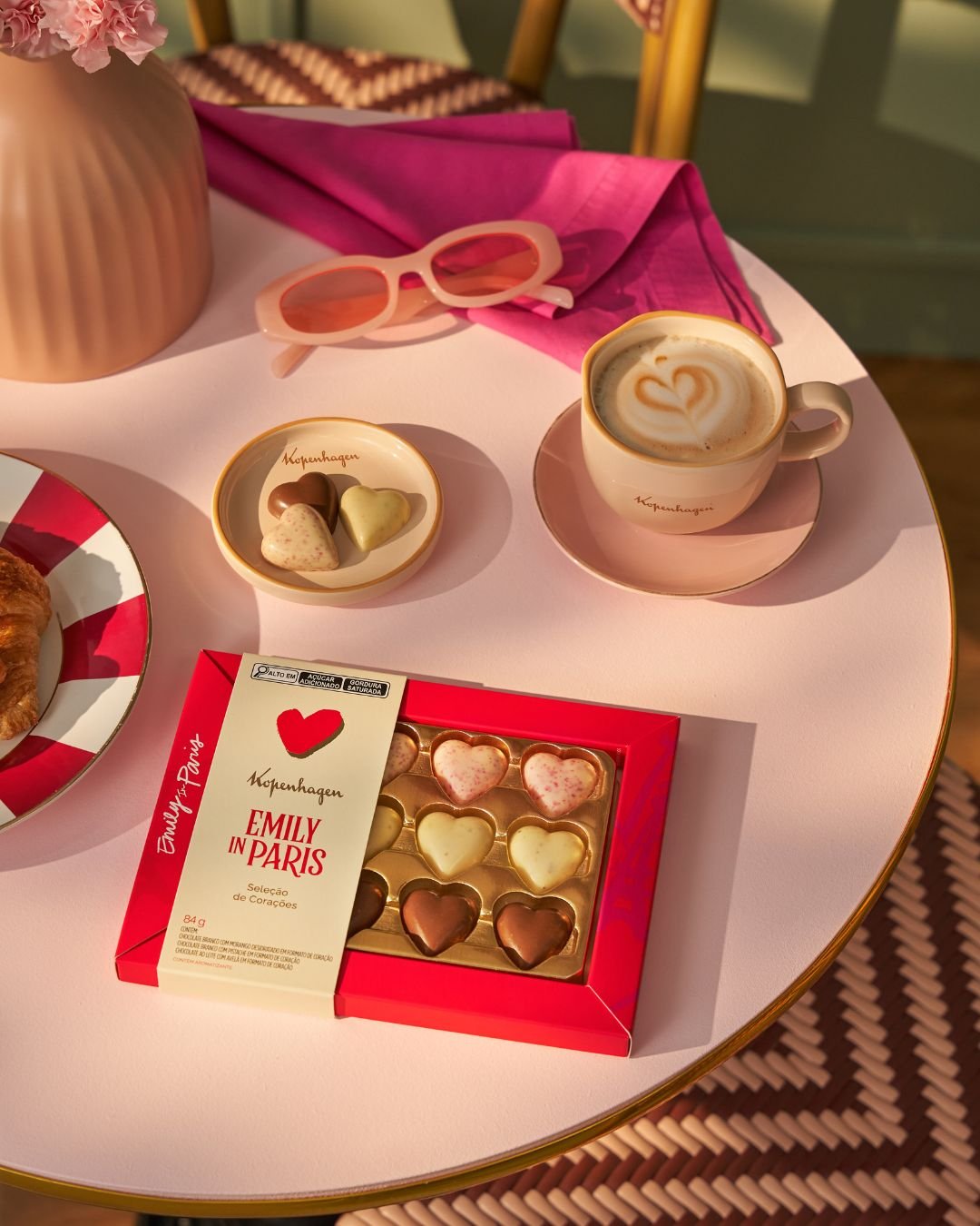 Chocolates em formato de coração ao lado de um café latte, óculos de sol e croissant em mesa elegante, evocando Dolce Far Niente e a estética de Emily In Paris