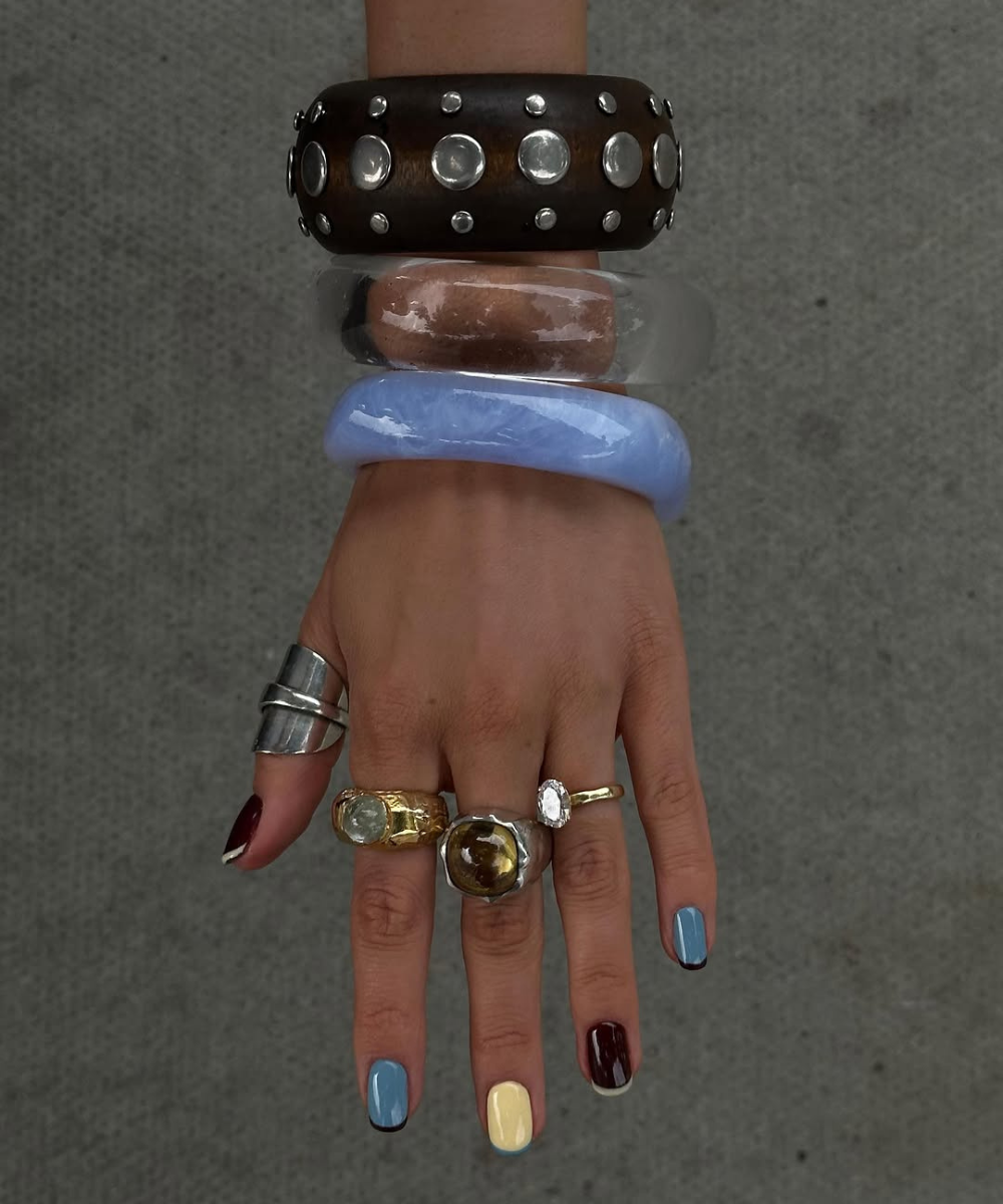 Pulseiras e anéis misturam prata e dourado, complementando unhas coloridas em azul, amarelo e vinho.