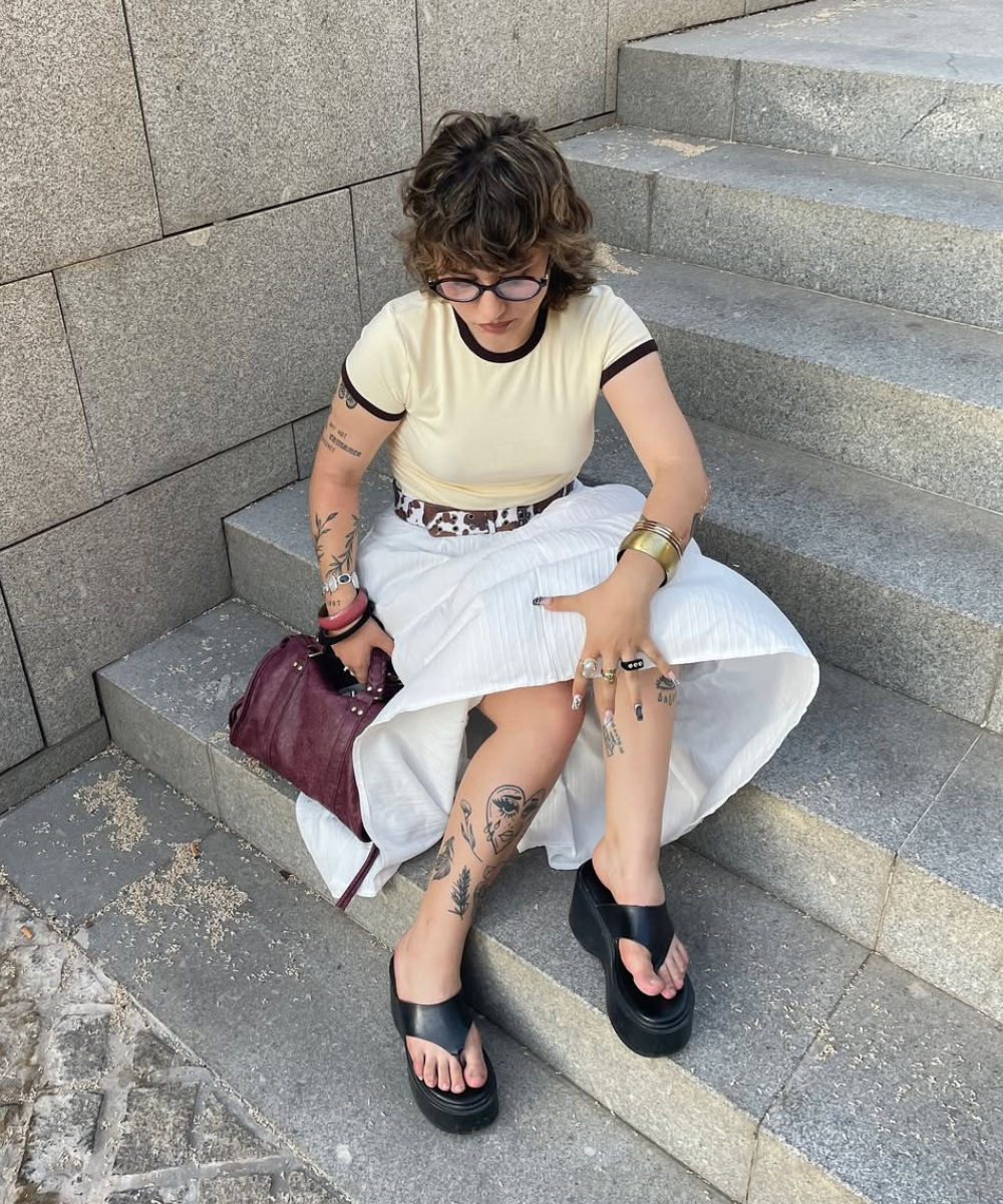 Pessoa sentada em escada de pedra, com tatuagens nos braços e pernas, usa sandálias pretas e segura bolsa vinho.