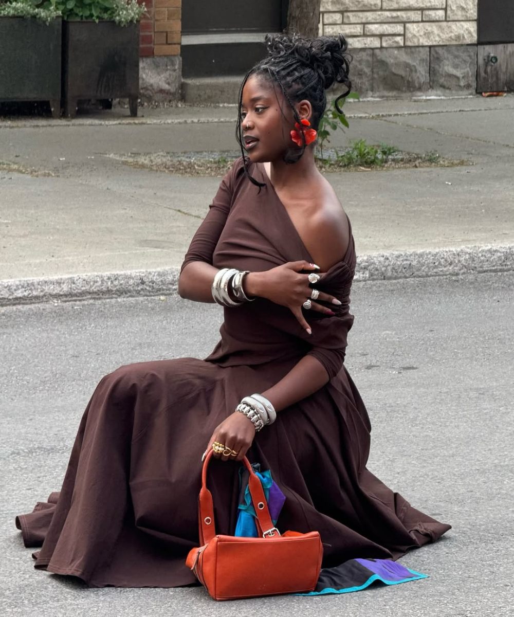 Mulher sentada na calçada usa vestidos e acessórios prateados, segurando bolsa laranja. Flor vermelha no cabelo.