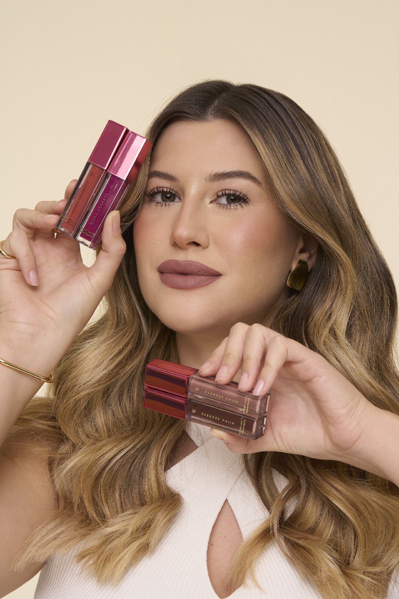 Mulher segurando batons líquidos Niina Secrets Neutrals com cores variadas e tampa rosa, exibindo maquiagem elegante.
