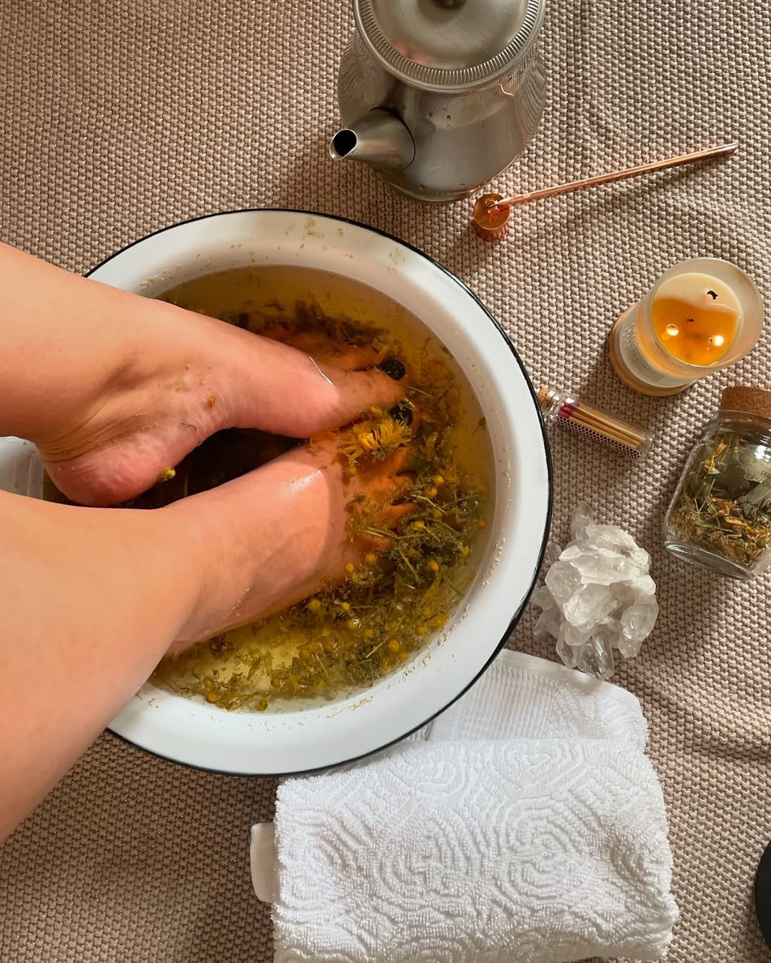 Pés em uma bacia de escalda-pés com ervas, ao lado de vela, chaleira e toalha.