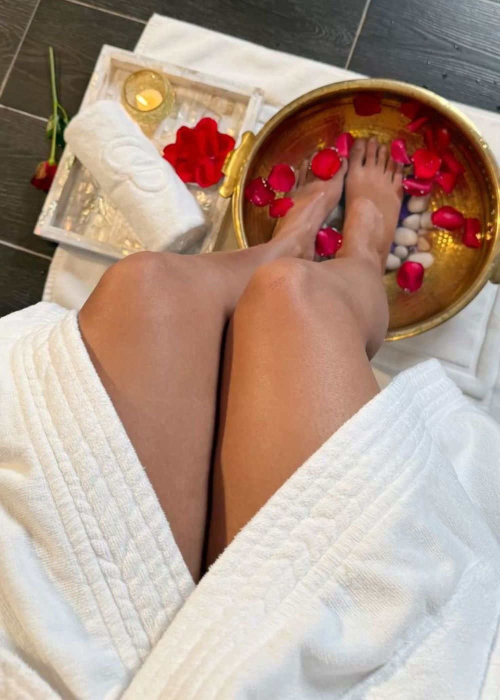 Pés mergulhados em bacia dourada com pétalas de rosa, acompanhados de toalha e vela. Escalda-pés relaxante.