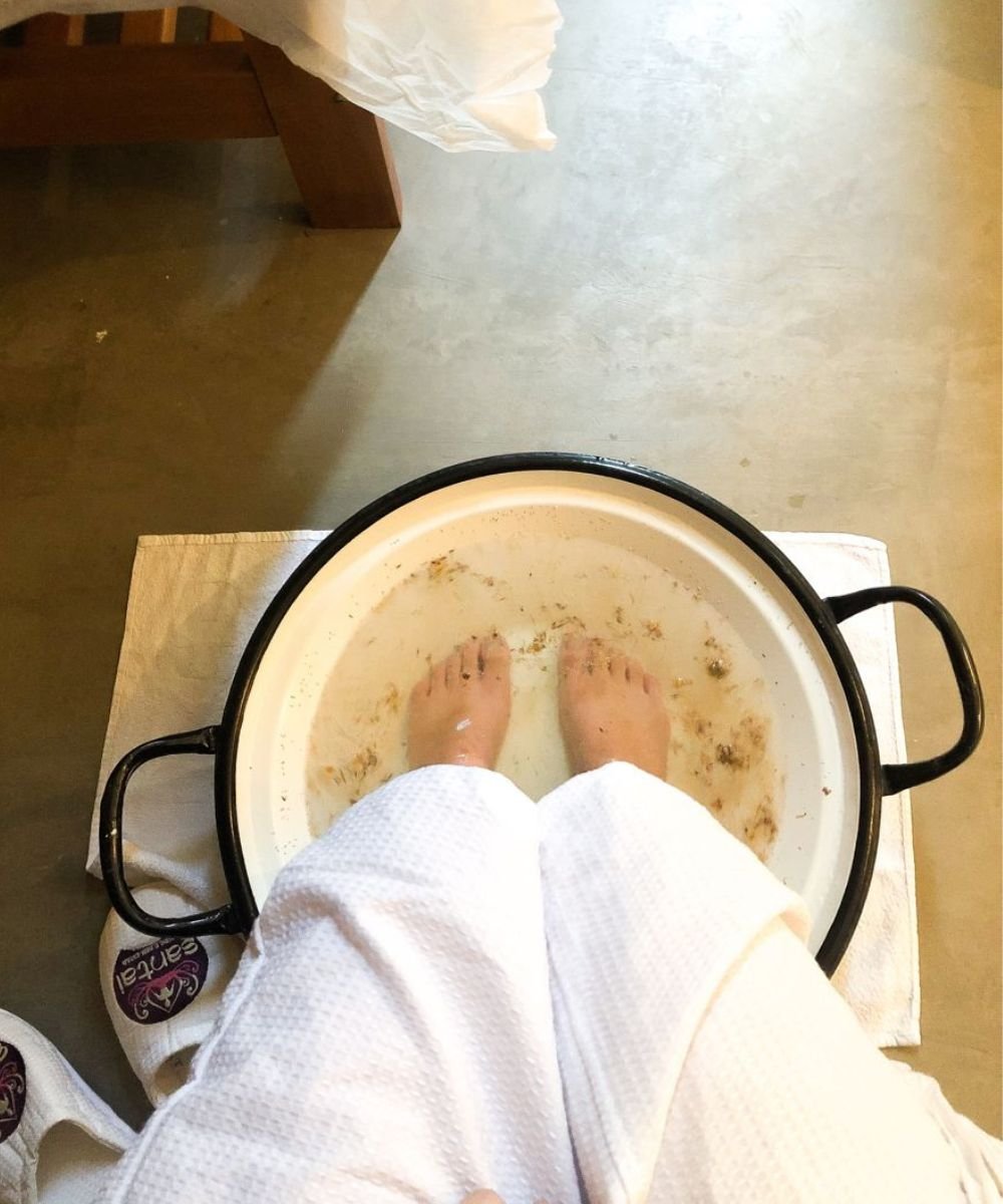 Pés relaxam em escalda-pés com água morna e ervas, enquanto pessoa usa roupão branco.