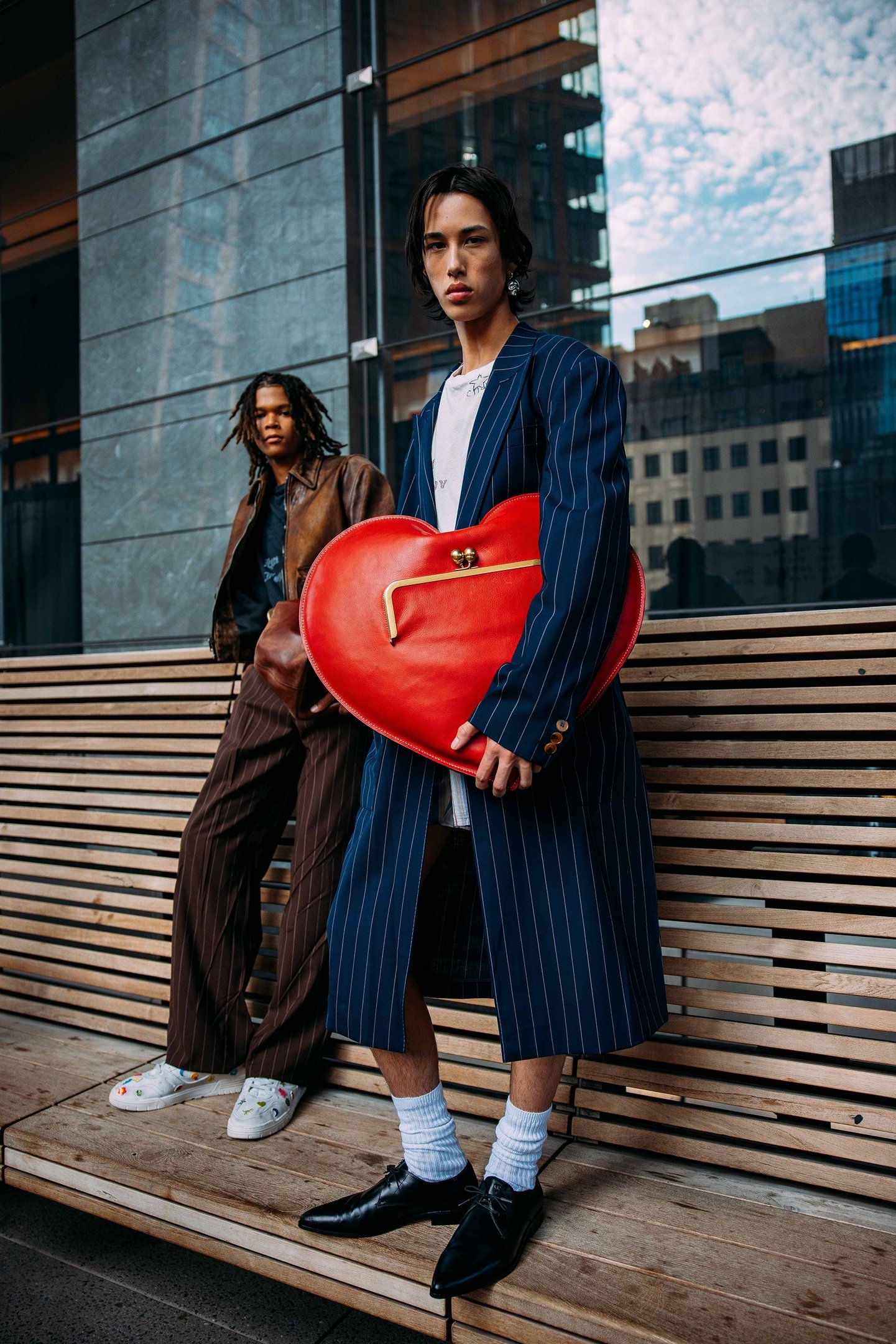 Homens usando roupas estilosas em espaço urbano, destacando bolsa vermelha em forma de coração.