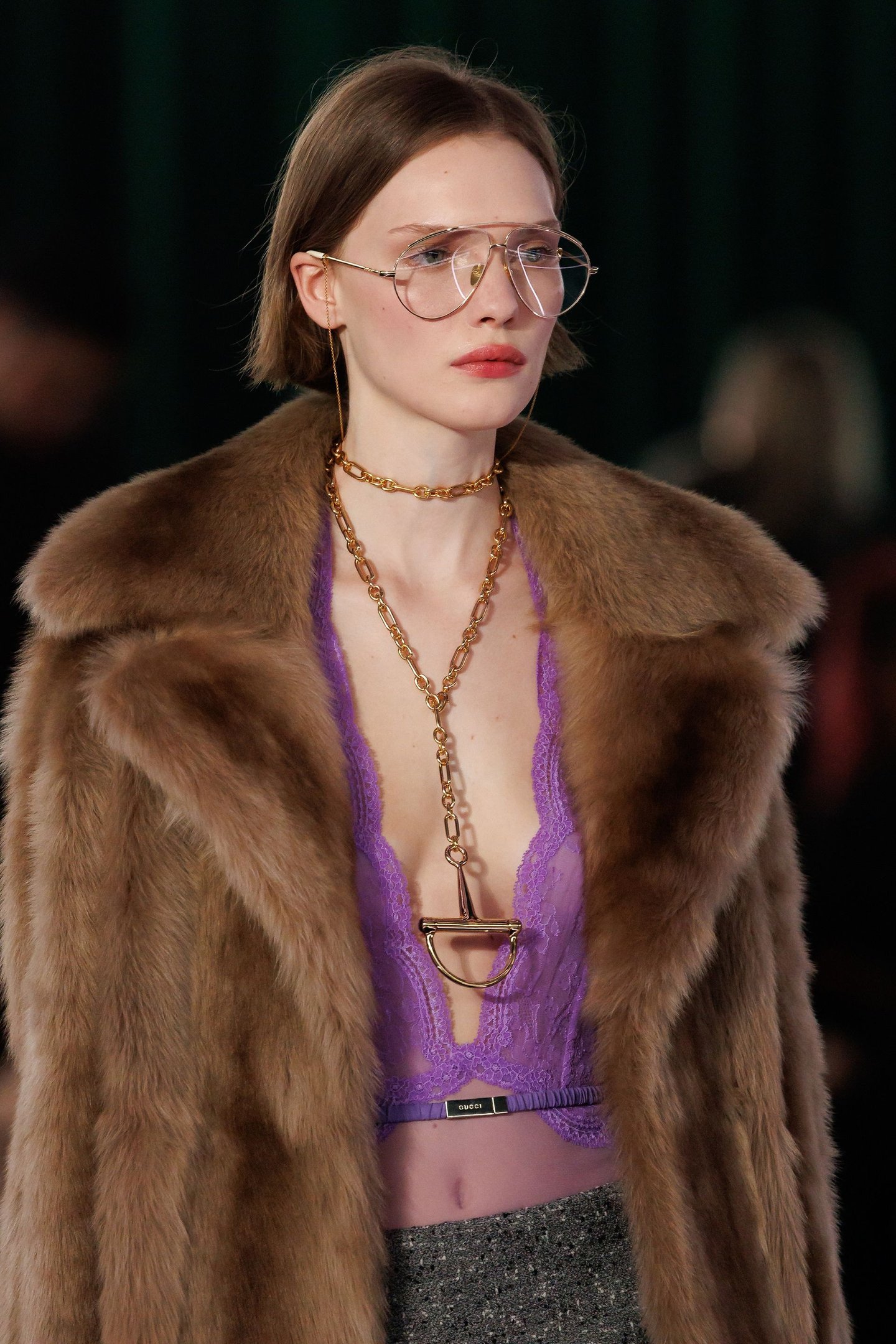 Parece haver um foco em moda durante o mês fashion: modelo em casaco marrom, top roxo de renda, óculos grandes e colar dourado.