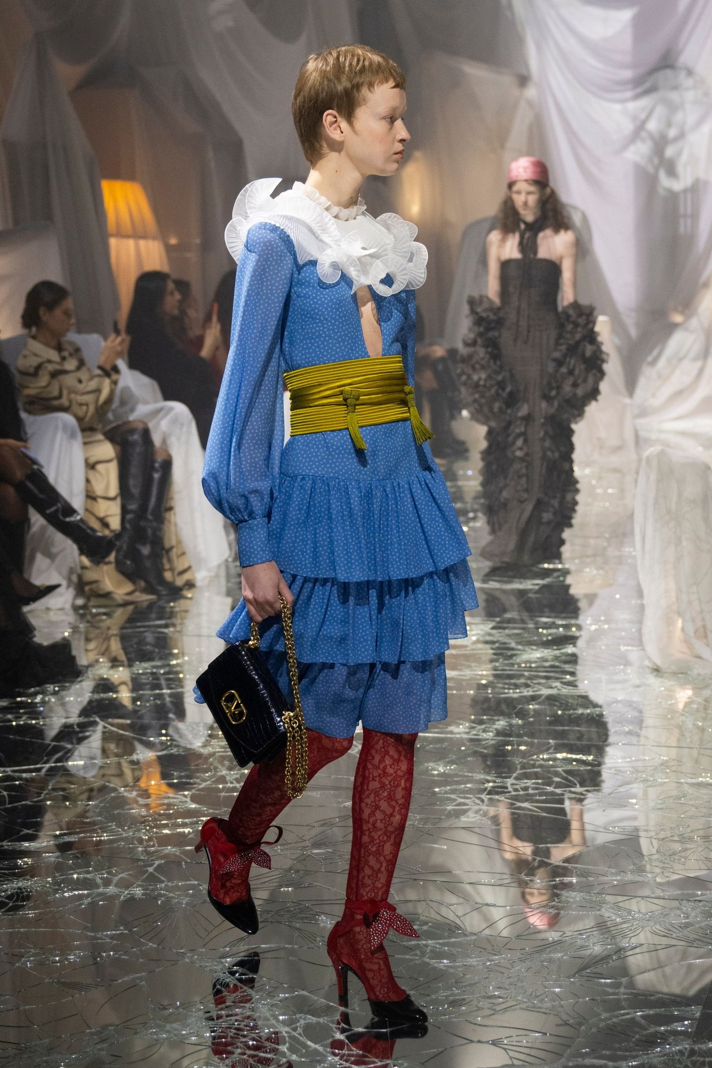 Desfile na passarela com vestido azul, babados brancos e cinto amarelo, destacando moda do mês fashion.