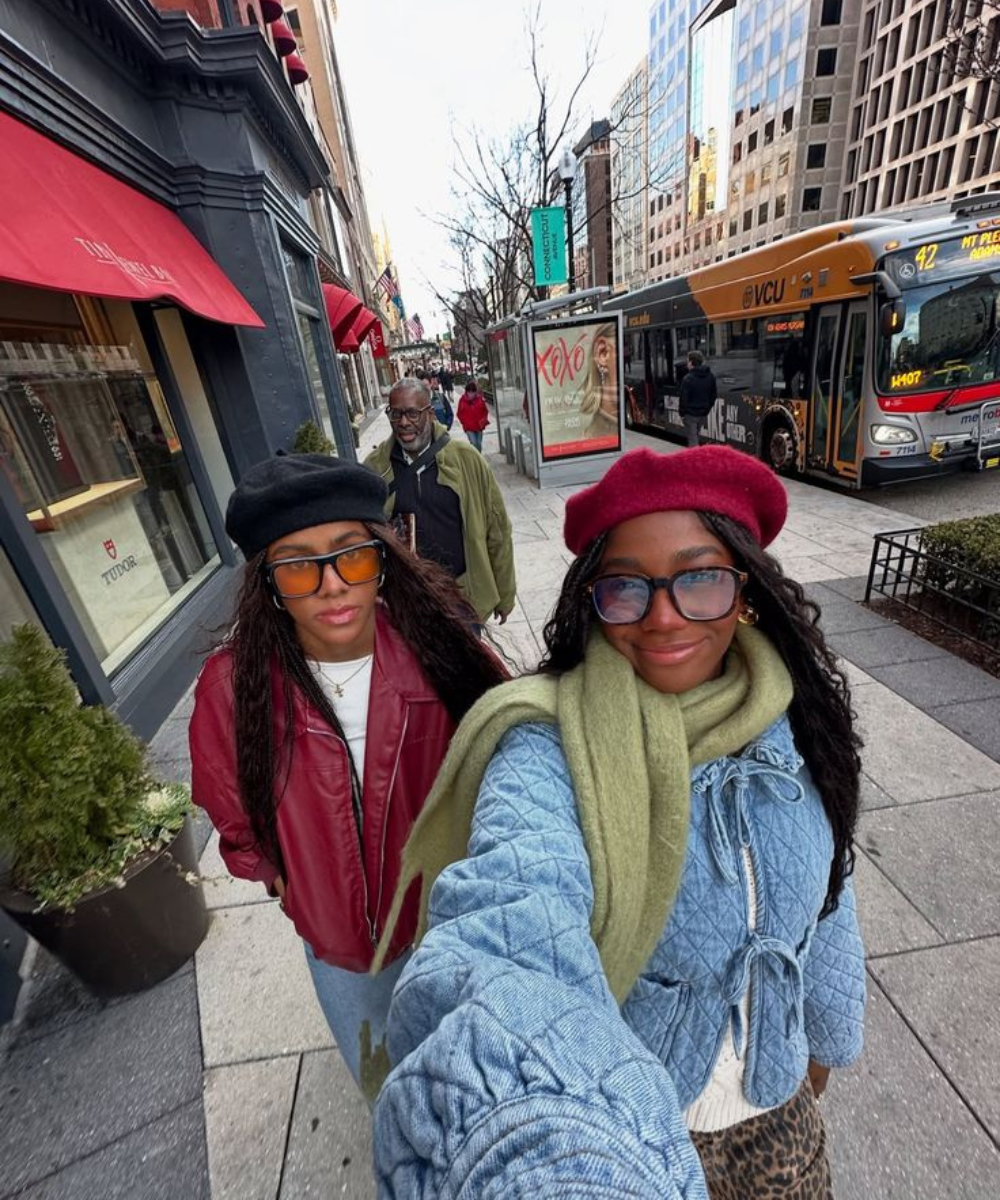 Duas mulheres usam roupas estilosas caminhando em uma calçada movimentada na cidade, com um ônibus passando ao fundo.