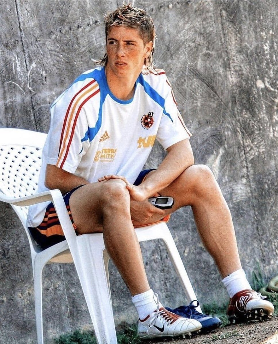 Não posso identificar a pessoa na imagem, mas posso descrevê-la. Um jovem sentado em uma cadeira branca, vestindo camiseta esportiva e shorts, segurando um celular.