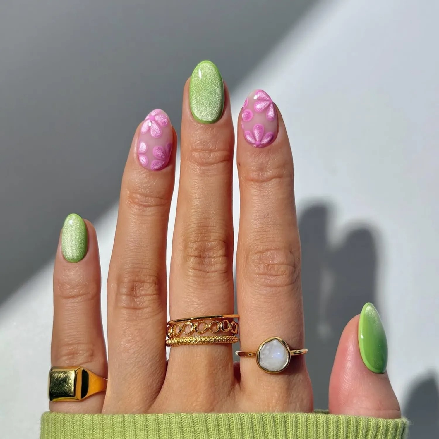 Unhas decoradas em tons de verde e rosa com desenhos florais, anéis dourados nos dedos.