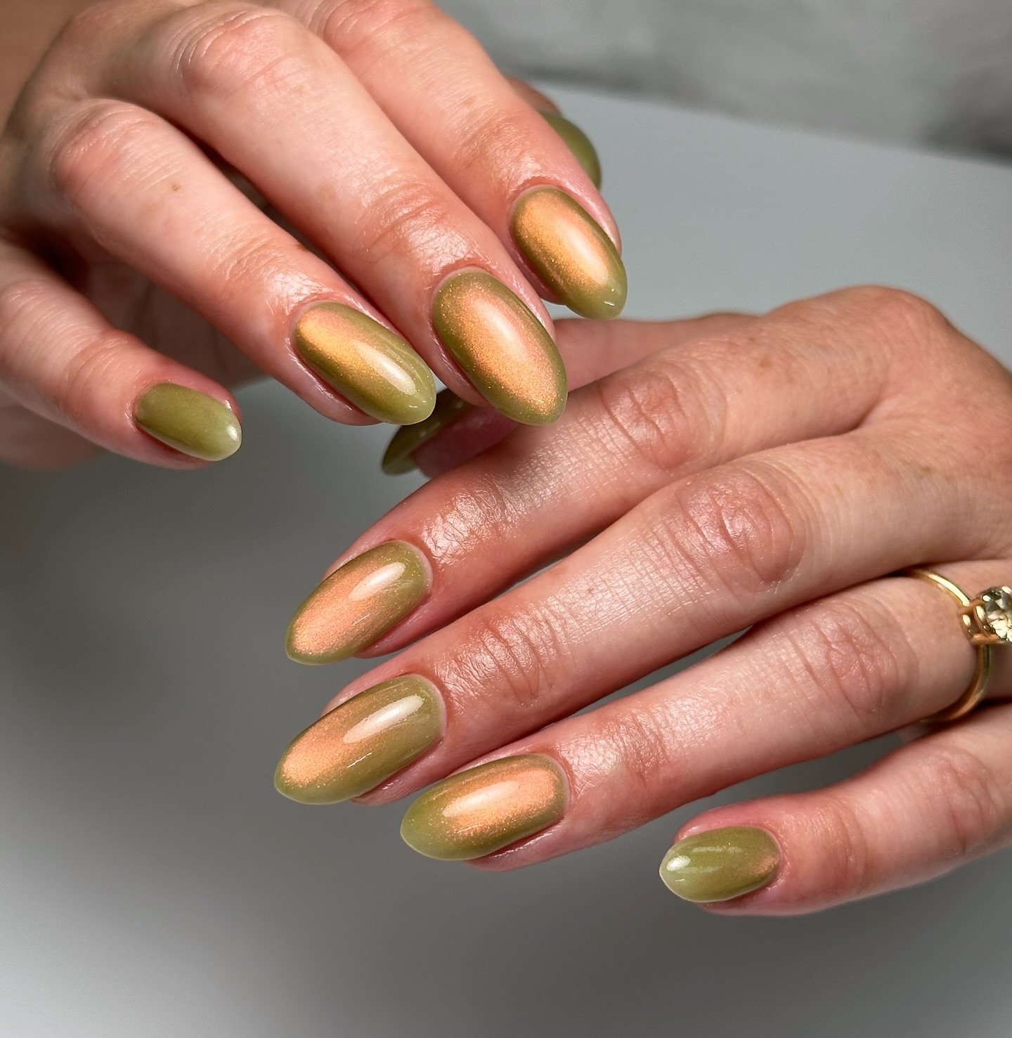 Unhas verdes com efeito metálico suave, estilo guava girl, em mãos entrelaçadas e anel dourado.