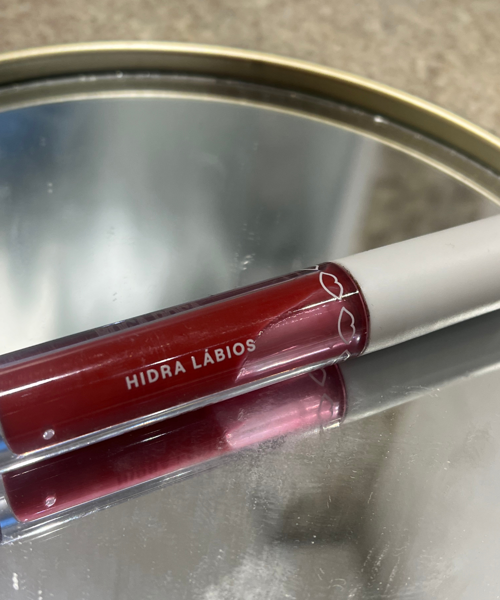 Brilho labial vermelho Hidra Lábios da marca Boca Rosa sobre uma superfície espelhada.