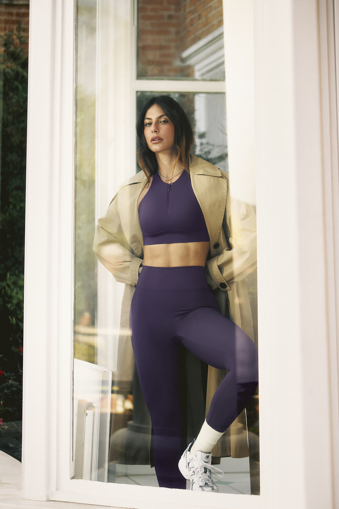 Pessoa vestindo conjunto esportivo roxo e casaco bege, perto de uma janela. Cenário sugere relaxamento ou moda em Hope Resort.