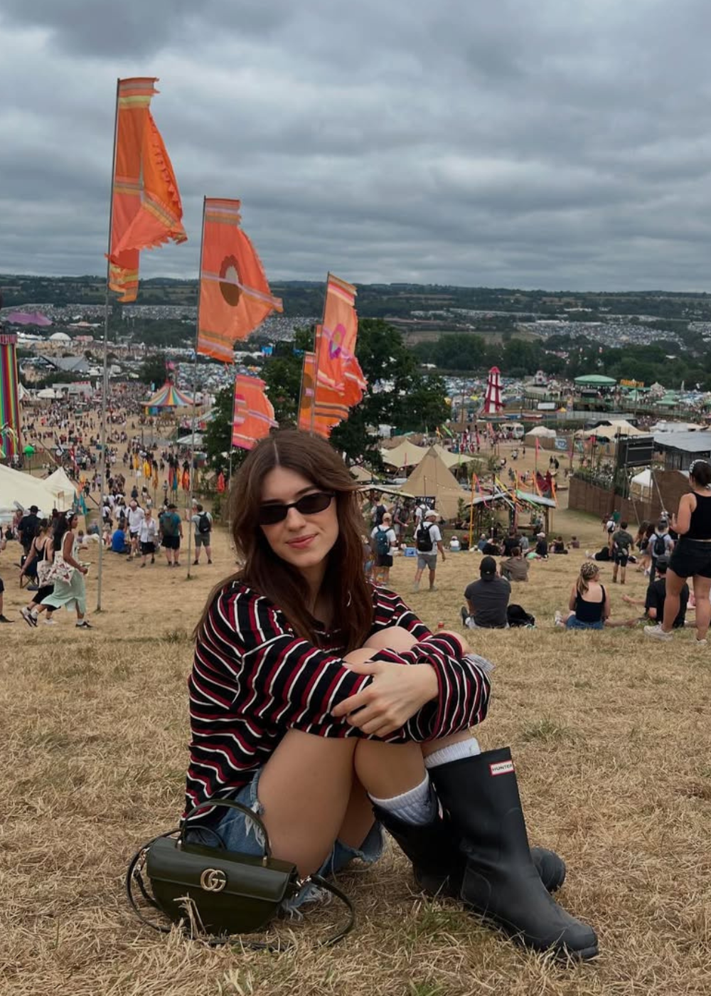 Jovem de óculos escuros e Hunter boots sentada na grama durante festival ao ar livre com bandeiras coloridas ao fundo.