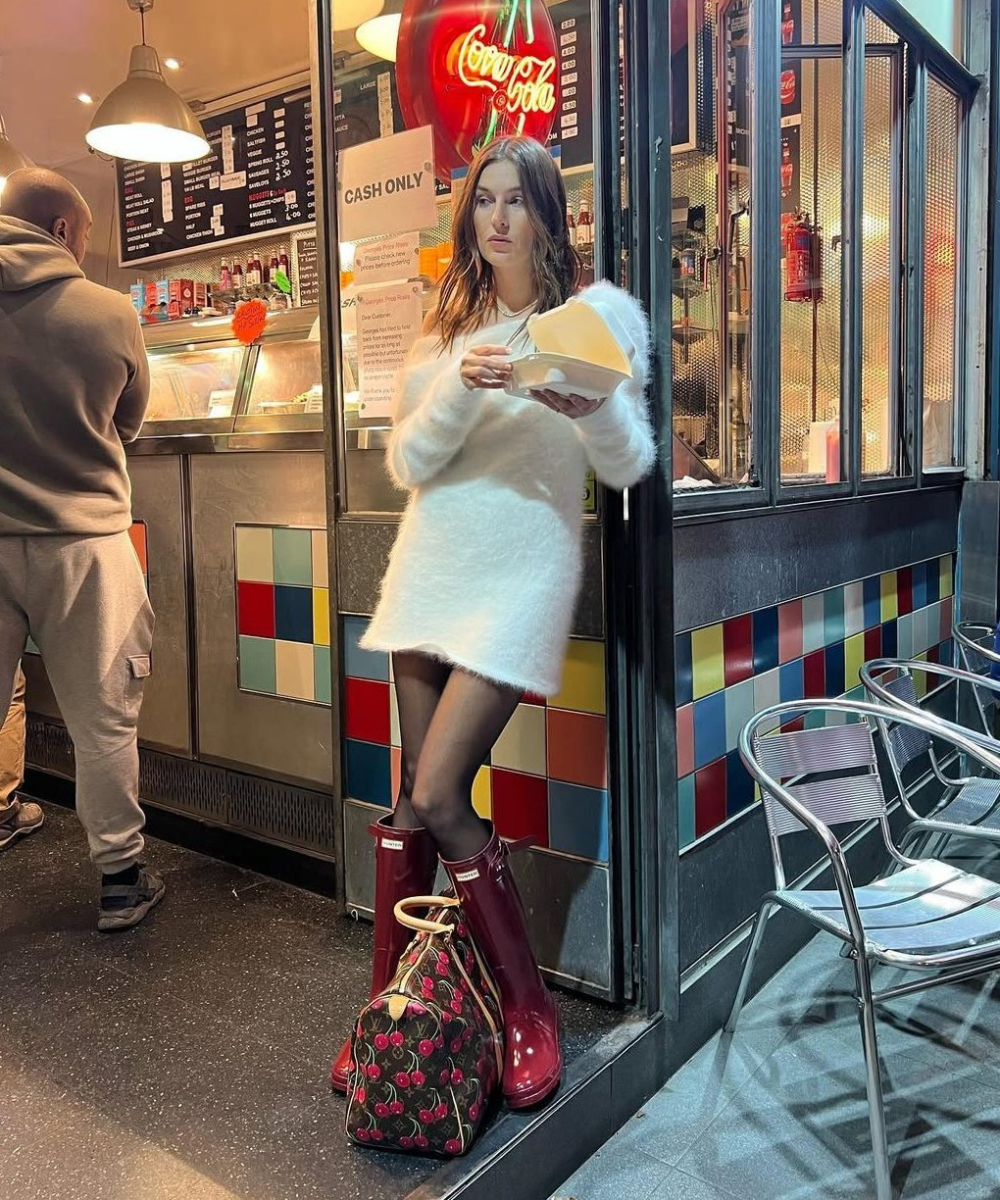 Mulher com botas vermelhas em frente a lanchonete, segurando comida. Bolsa estampada ao seu lado.