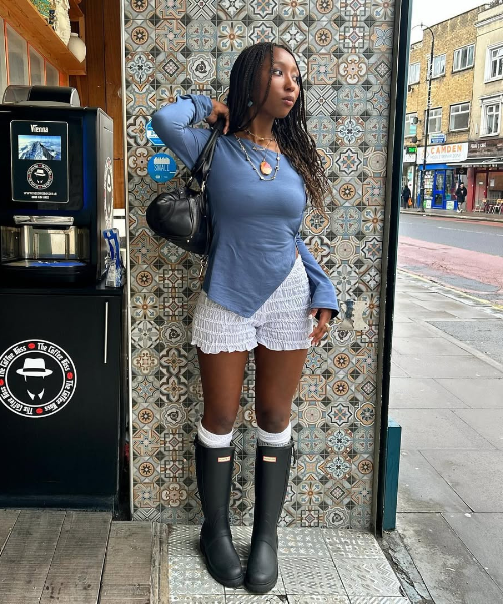 Pessoa com botas Hunter, camisa azul e shorts brancos em frente a parede de azulejos decorativos.