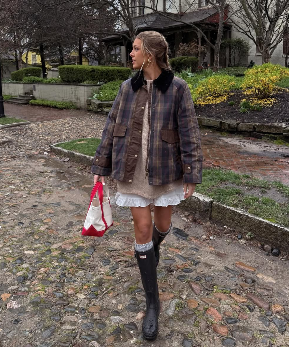 Mulher usando casaco xadrez e botas de chuva pretas, caminhando em rua de paralelepípedo com bolsa rosa e branca.
