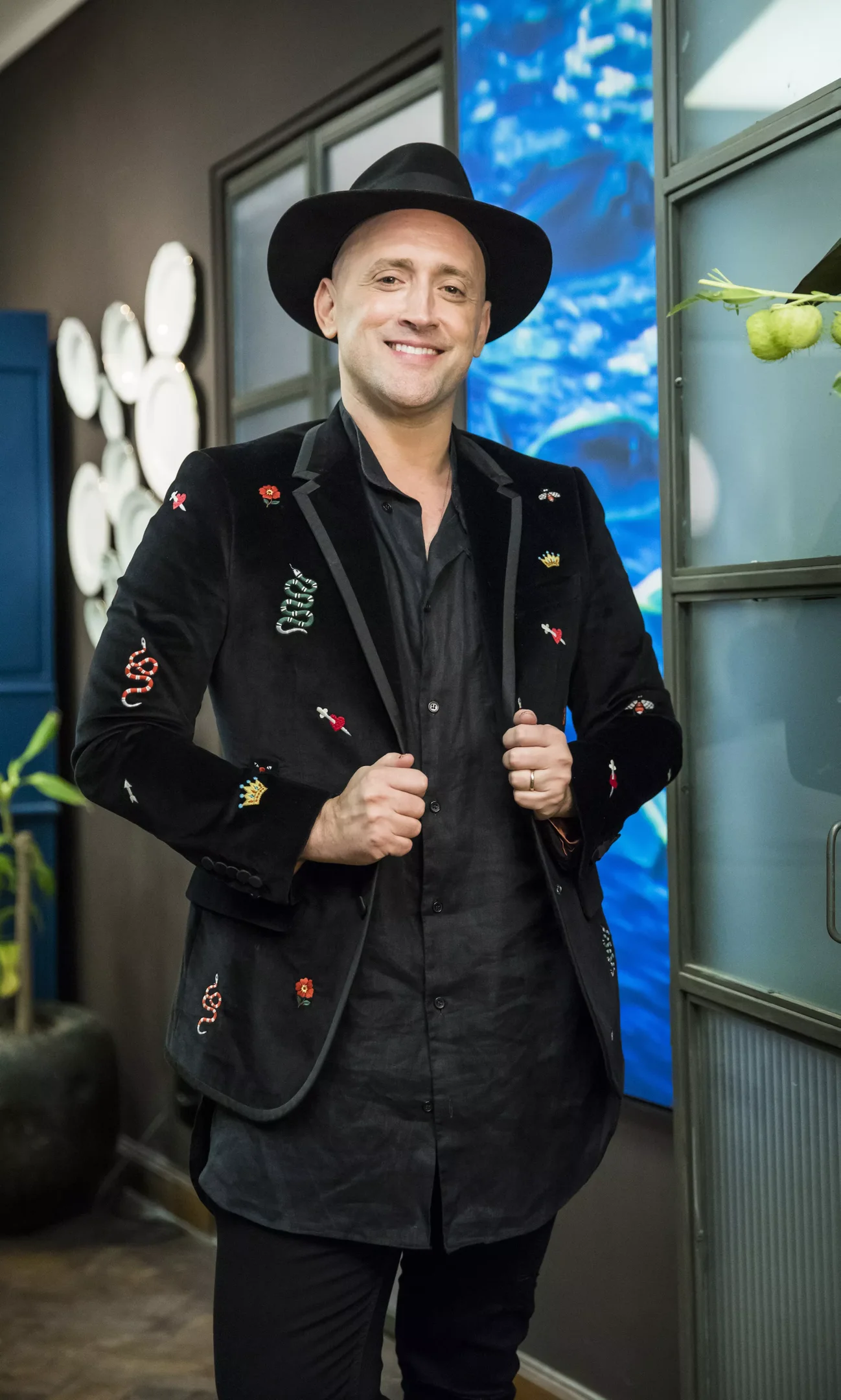 Leilão Paulo Gustavo sorridente veste terno bordado e chapéu, em ambiente interno.