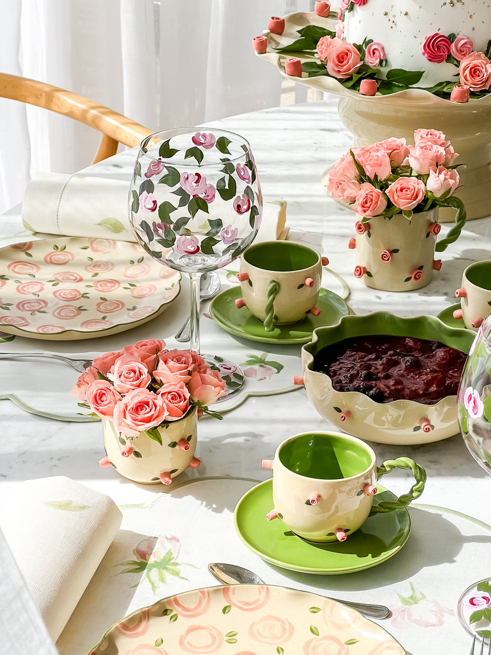 Mesa decorada com louças florais, copo e prato com rosas, bule e xícara em tons de verde. Flores e bolo ao fundo.