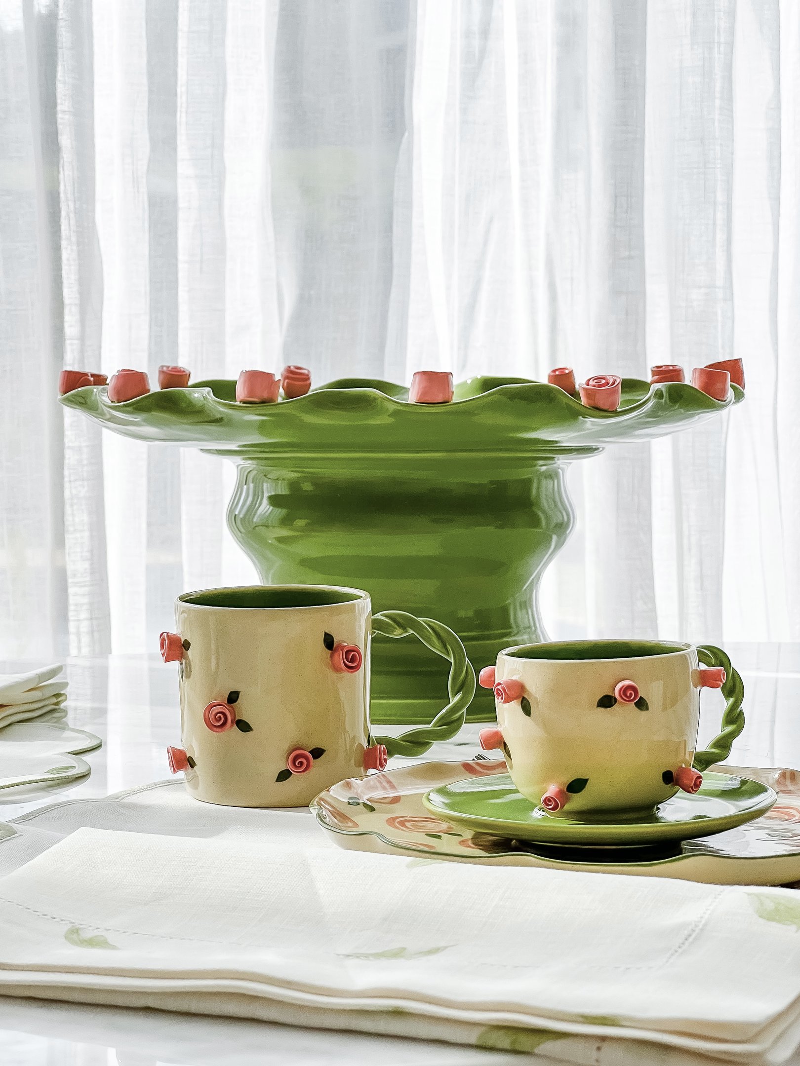 Conjunto de louça decorado com rosas vermelhas e verdes sobre mesa clara, design elegante de Marô Antunes.