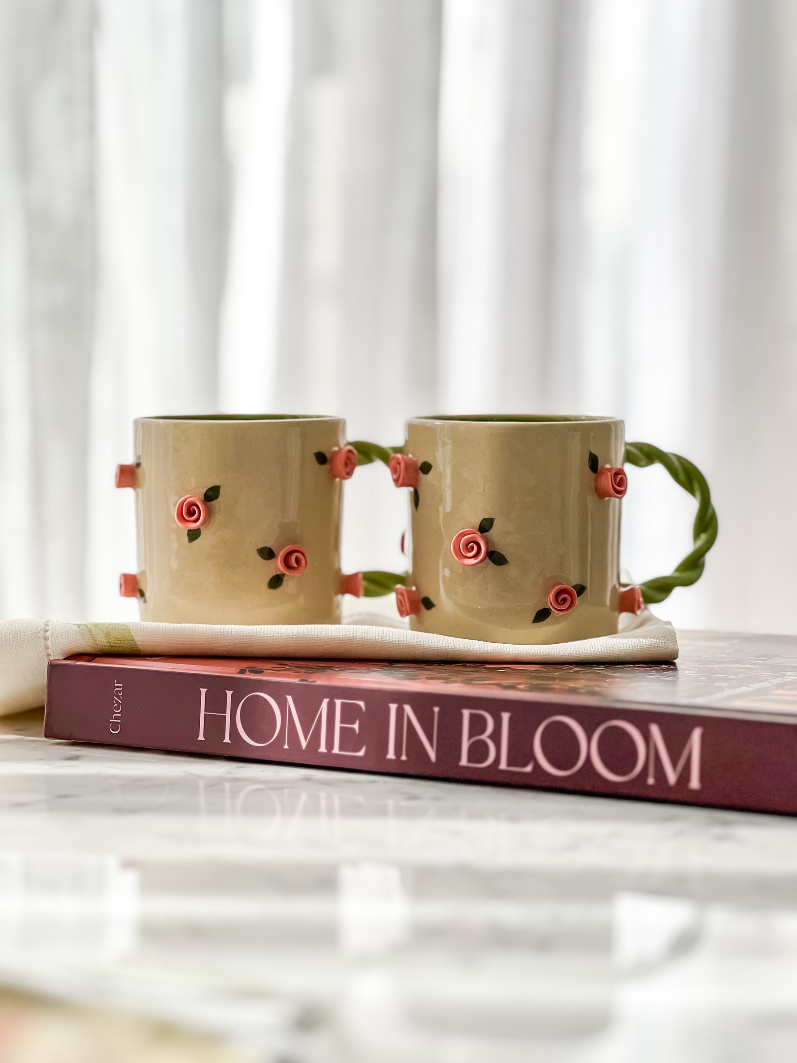 Duas canecas decoradas com rosas em fundo neutro, ao lado de um livro intitulado