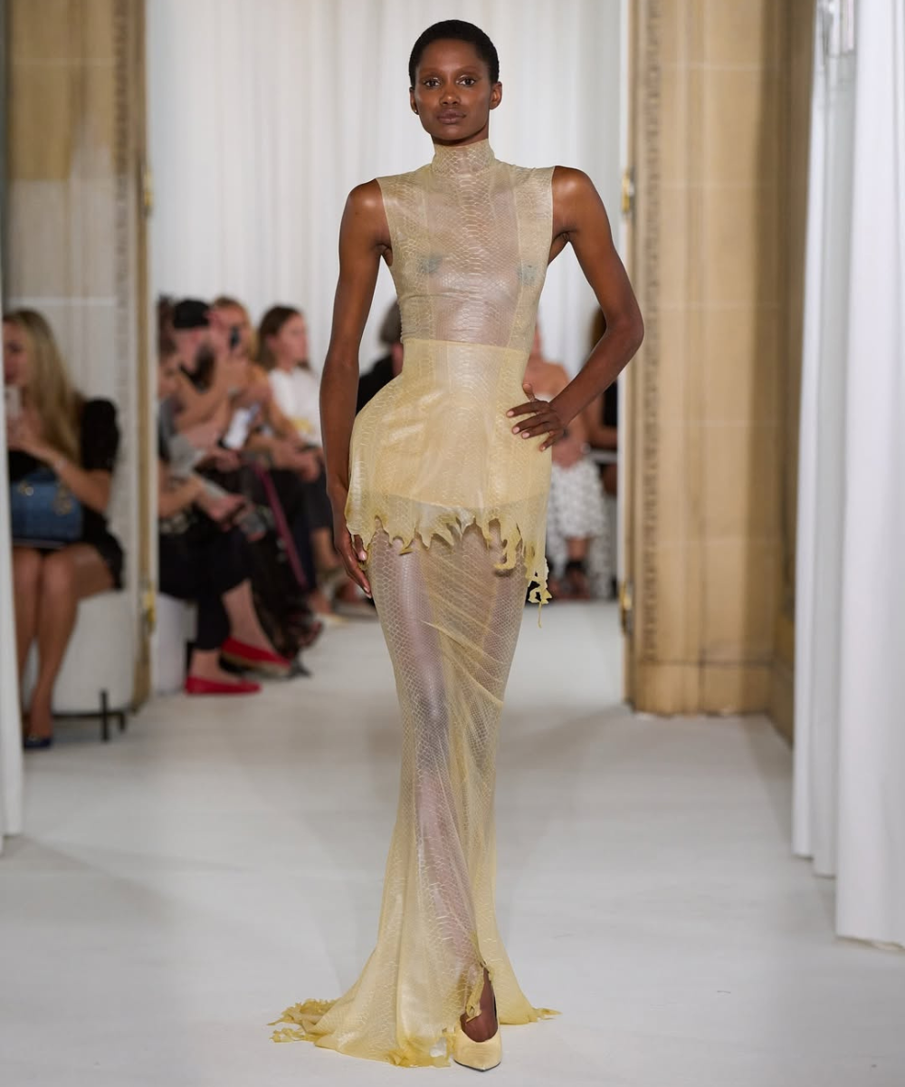 Modelo veste vestido transparente e dourado em desfile, com platéia ao fundo. Schiaparelli é sugerido.