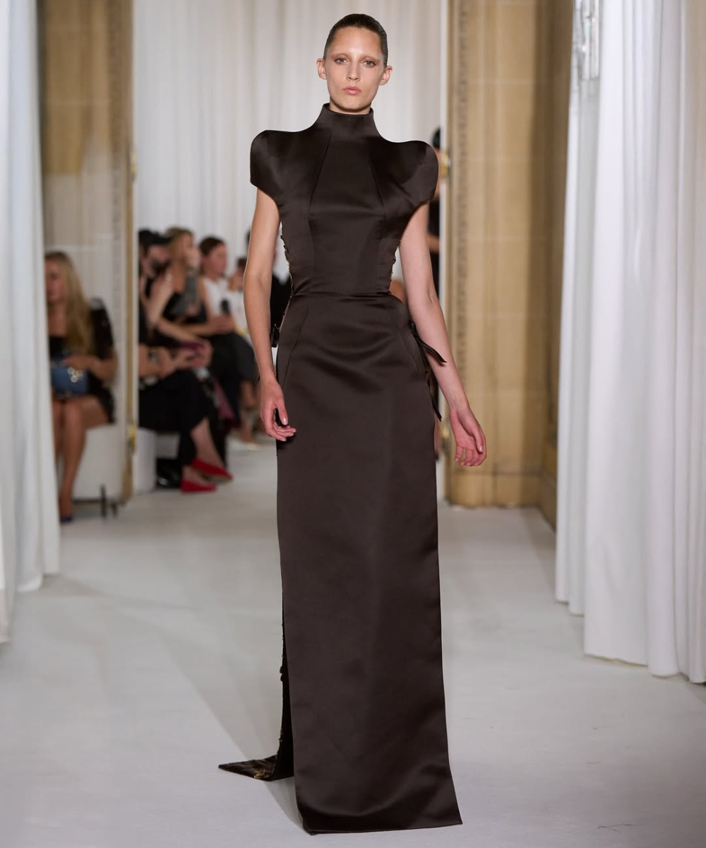 Modelo desfilando em vestido marrom escuro sofisticado, com ombros estruturados e gola alta, inspirado por Schiaparelli.