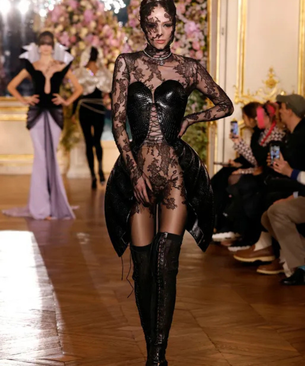 Desfile de moda com modelo em vestido de renda preta e corpete estruturado. Público assiste atento.