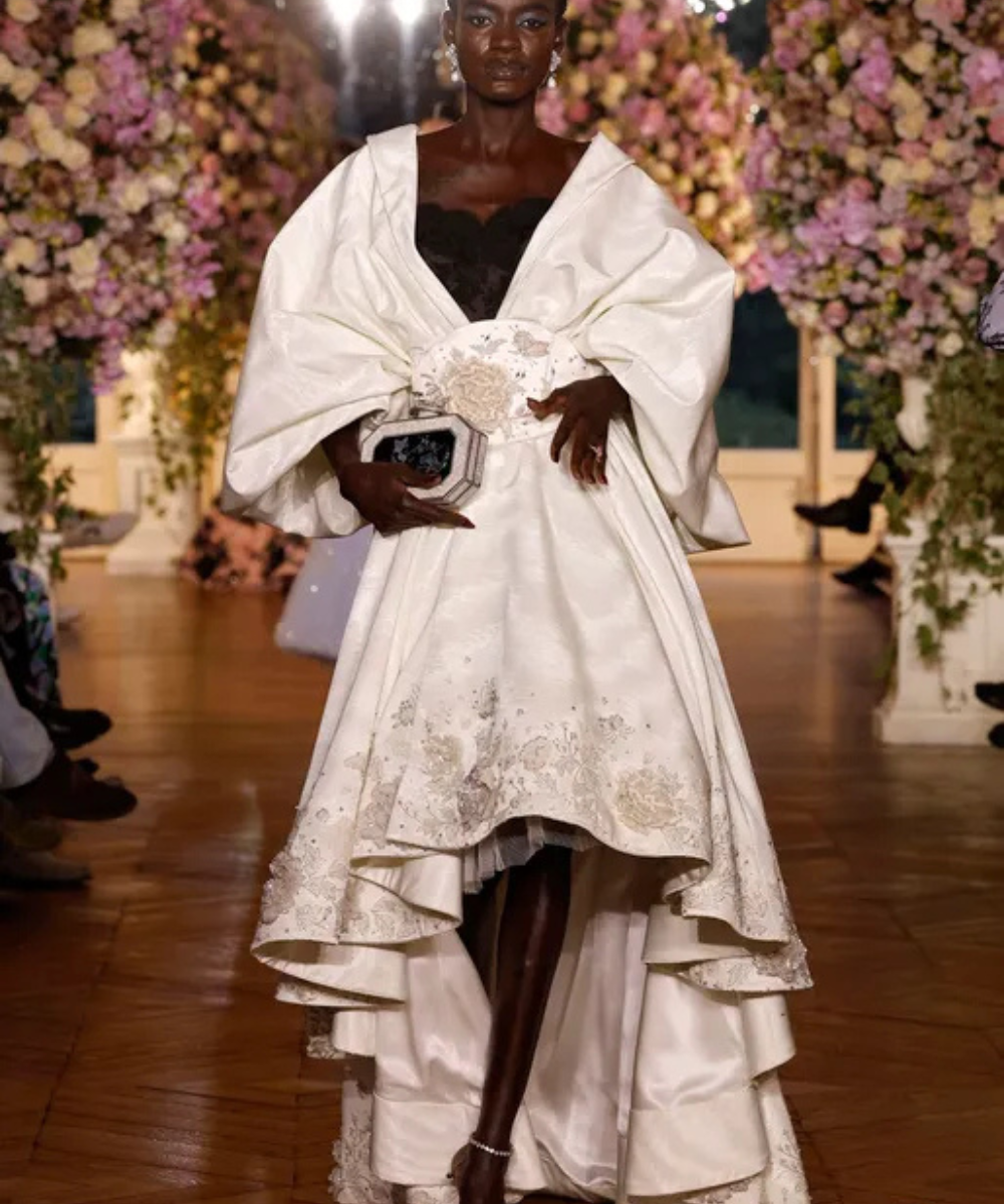 Modelo em vestido branco luxuoso com detalhes florais, desfilando em ambiente decorado com flores. Schiaparelli.