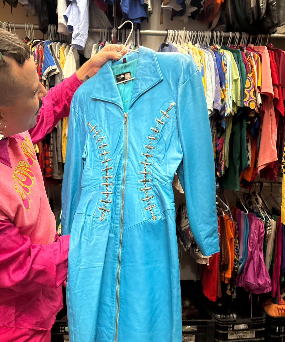 Pessoa segura macacão azul vivo com zíperes prateados, rodeado por roupas coloridas em loja vintage.