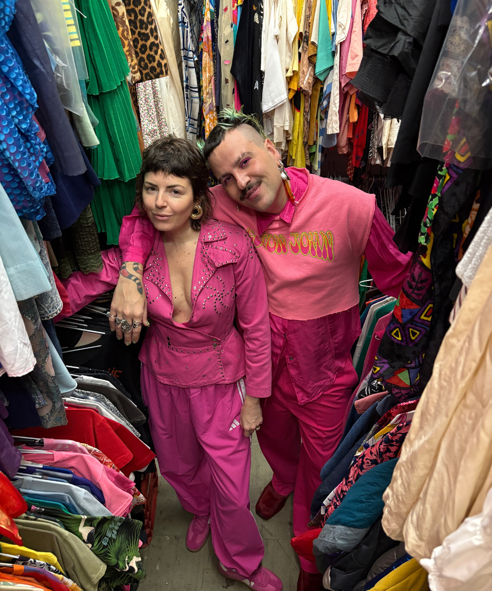 Duas pessoas em um brechó, vestindo roupas rosas chamativas, expressando estilo podre de chique.