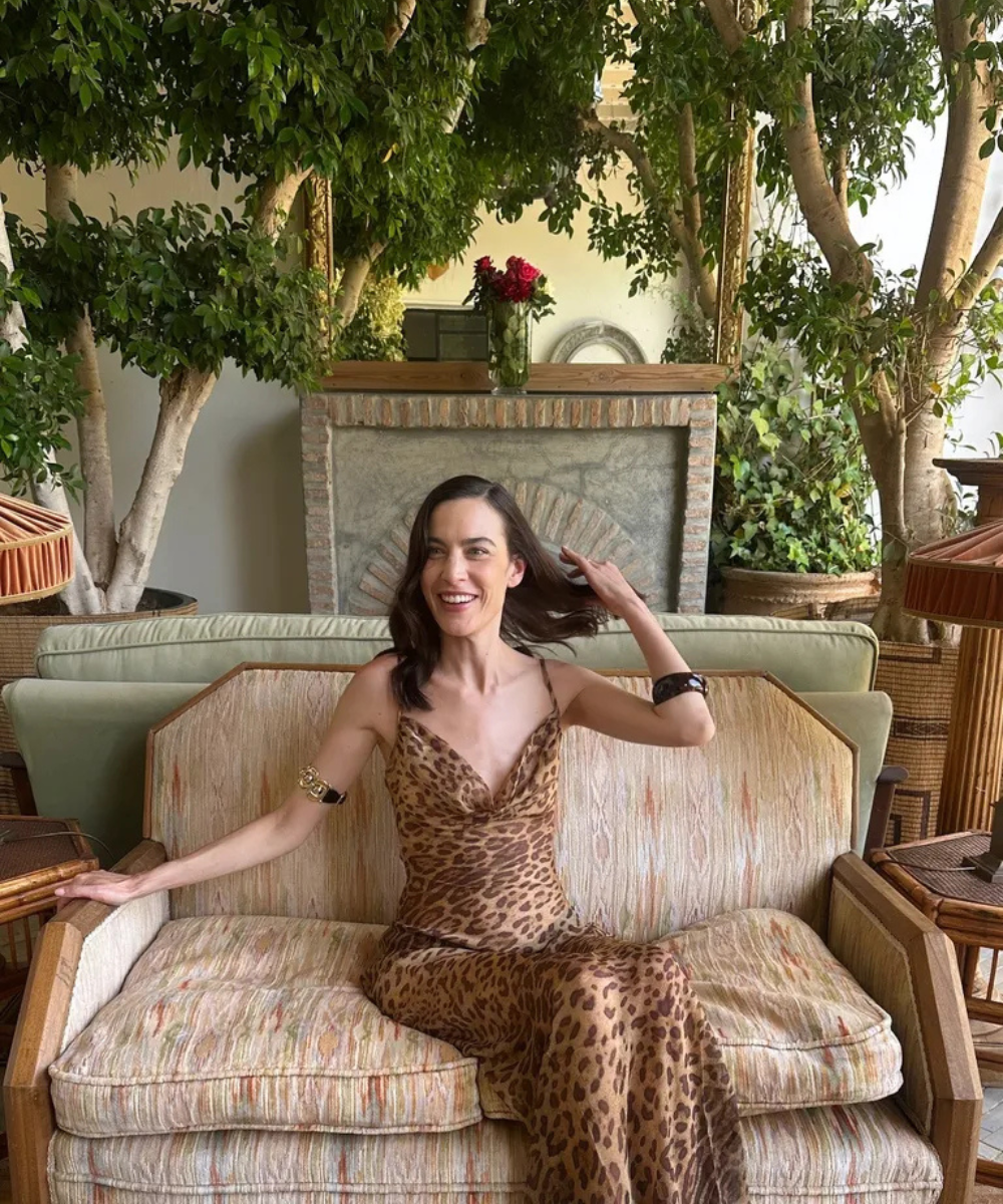 Mulher em vestido de estampa animal em sofá vintage, usa pulseira como acessório dos anos 80, rodeada por plantas.