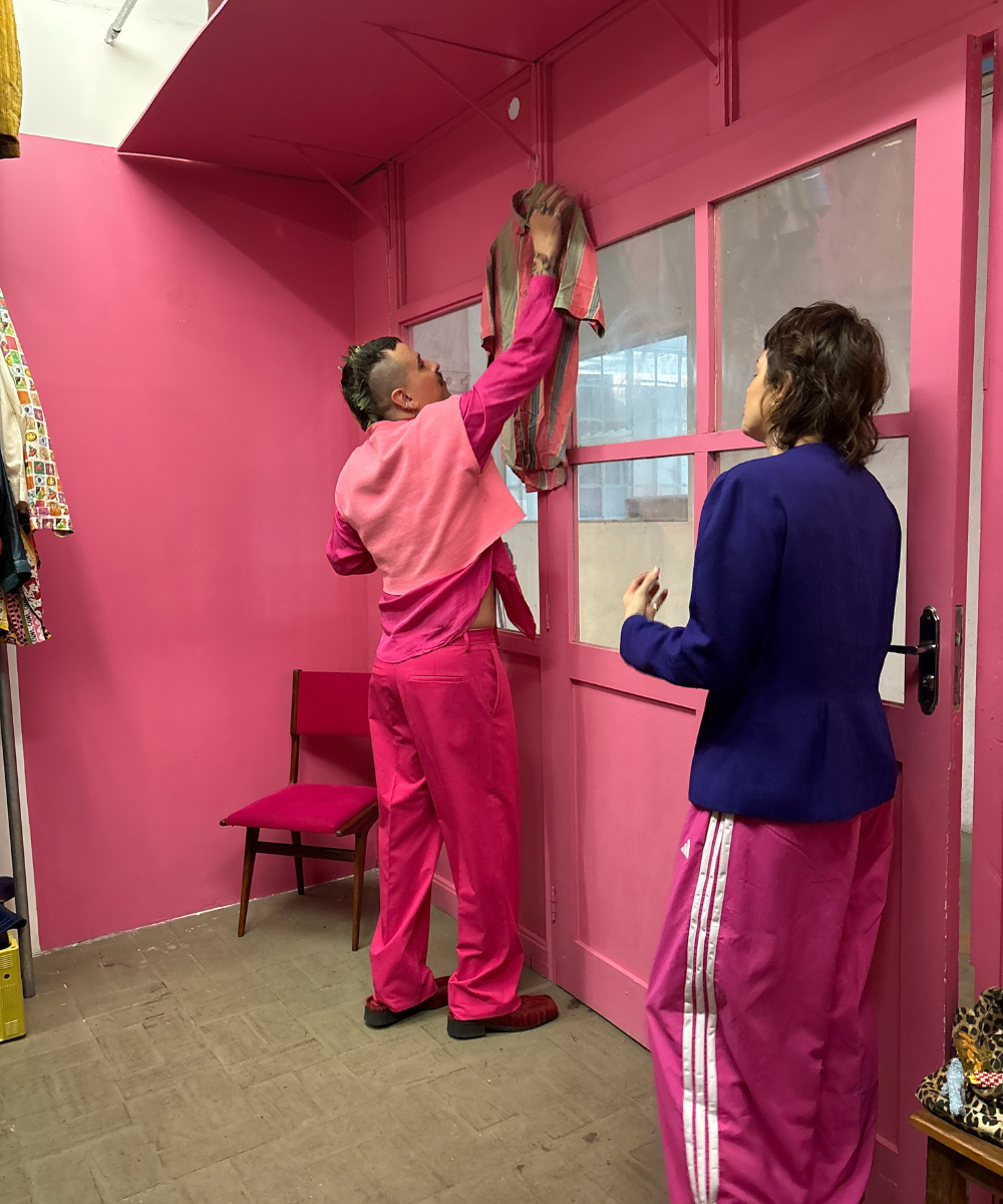 Duas pessoas em roupas coloridas em um ambiente dominado por um tom de rosa vibrante, organizando roupas. Podre de chique!