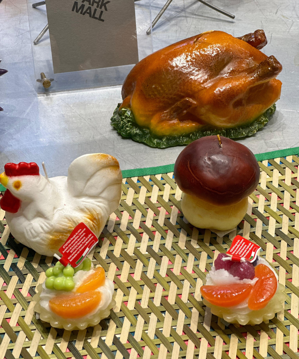Velas decorativas em forma de frango, cogumelo e tortinhas de frutas estão expostas sobre uma superfície trançada.