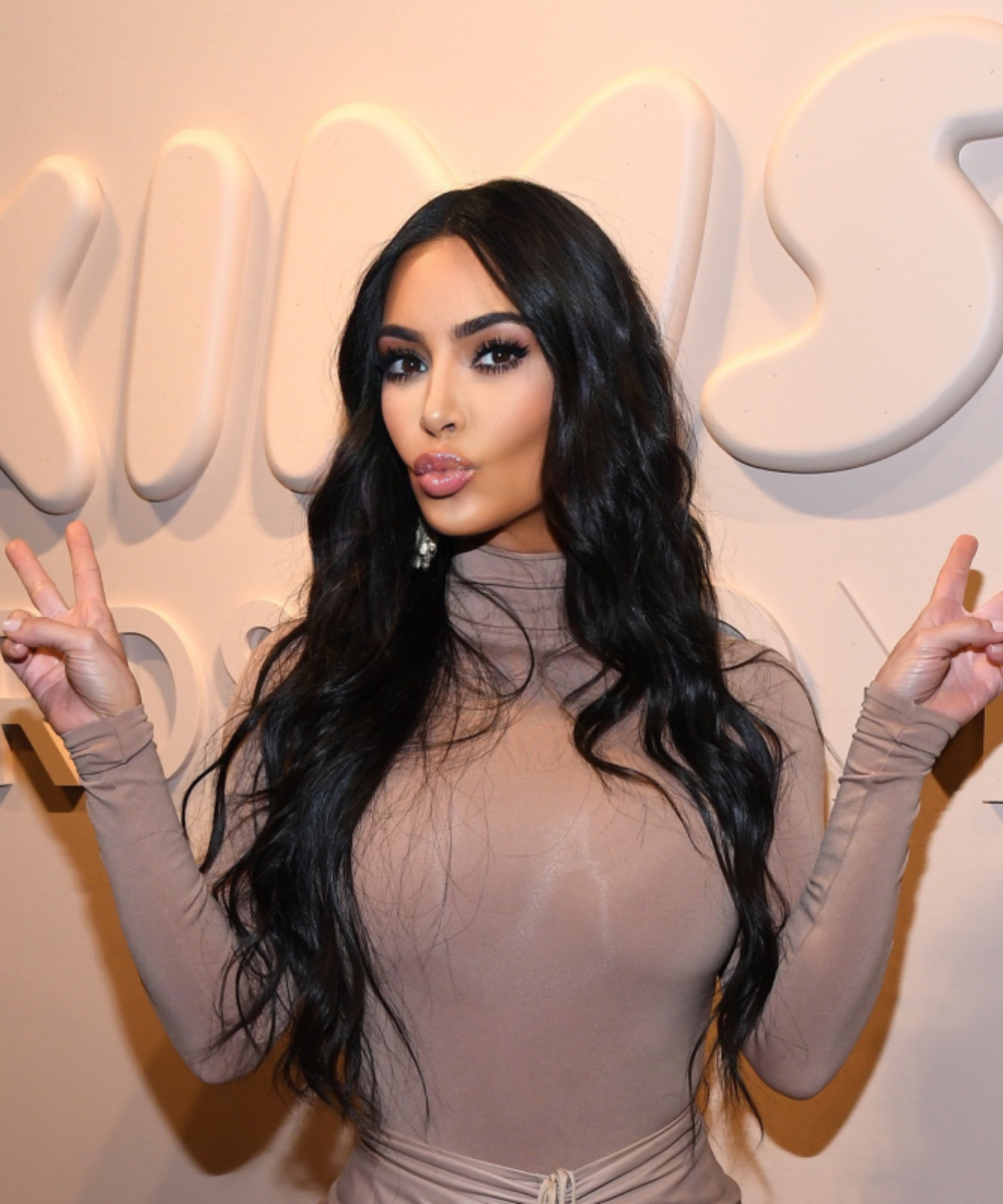 Mulher com cabelo longo e ondulado faz sinal de paz com as mãos. Ambiente relacionado a cuidados com a pele.