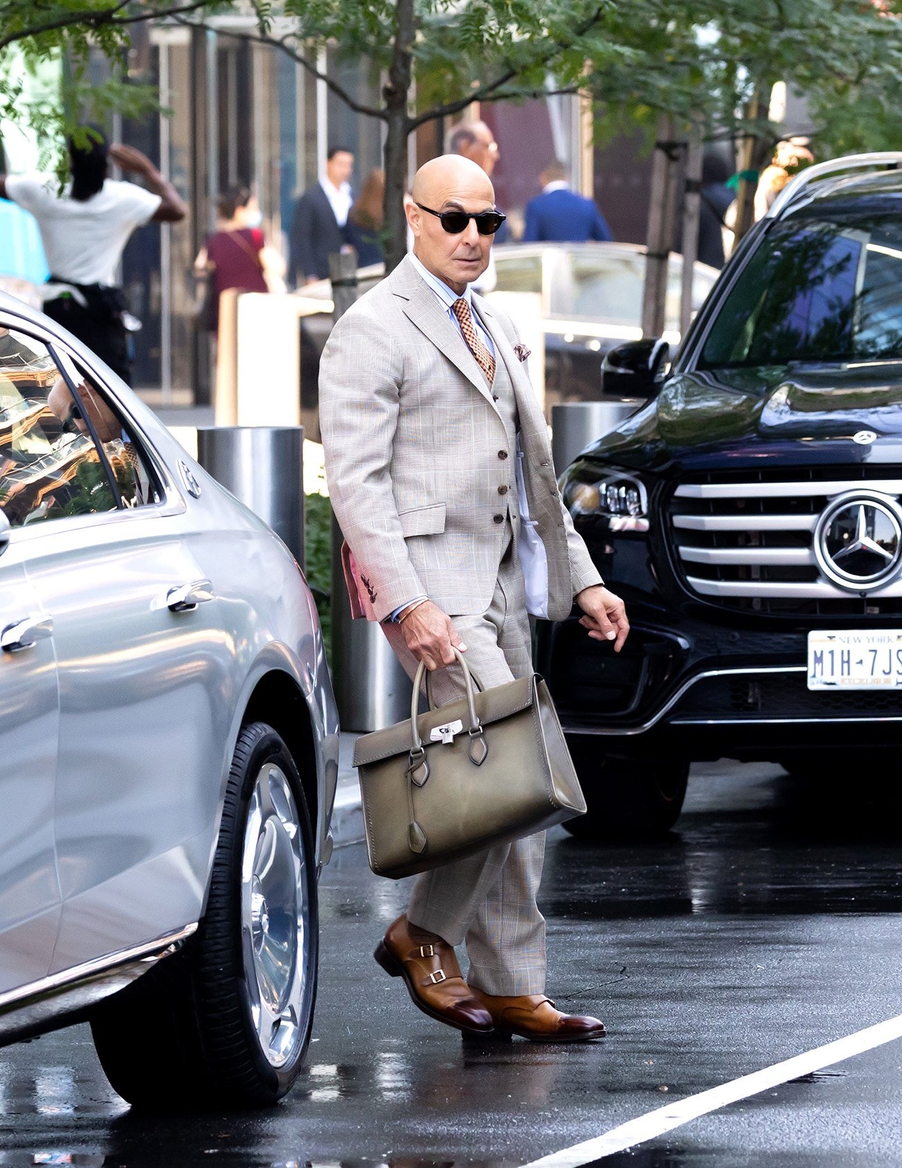 Homem de terno cinza e óculos escuros, carregando pasta sofisticada, em ambiente urbano.