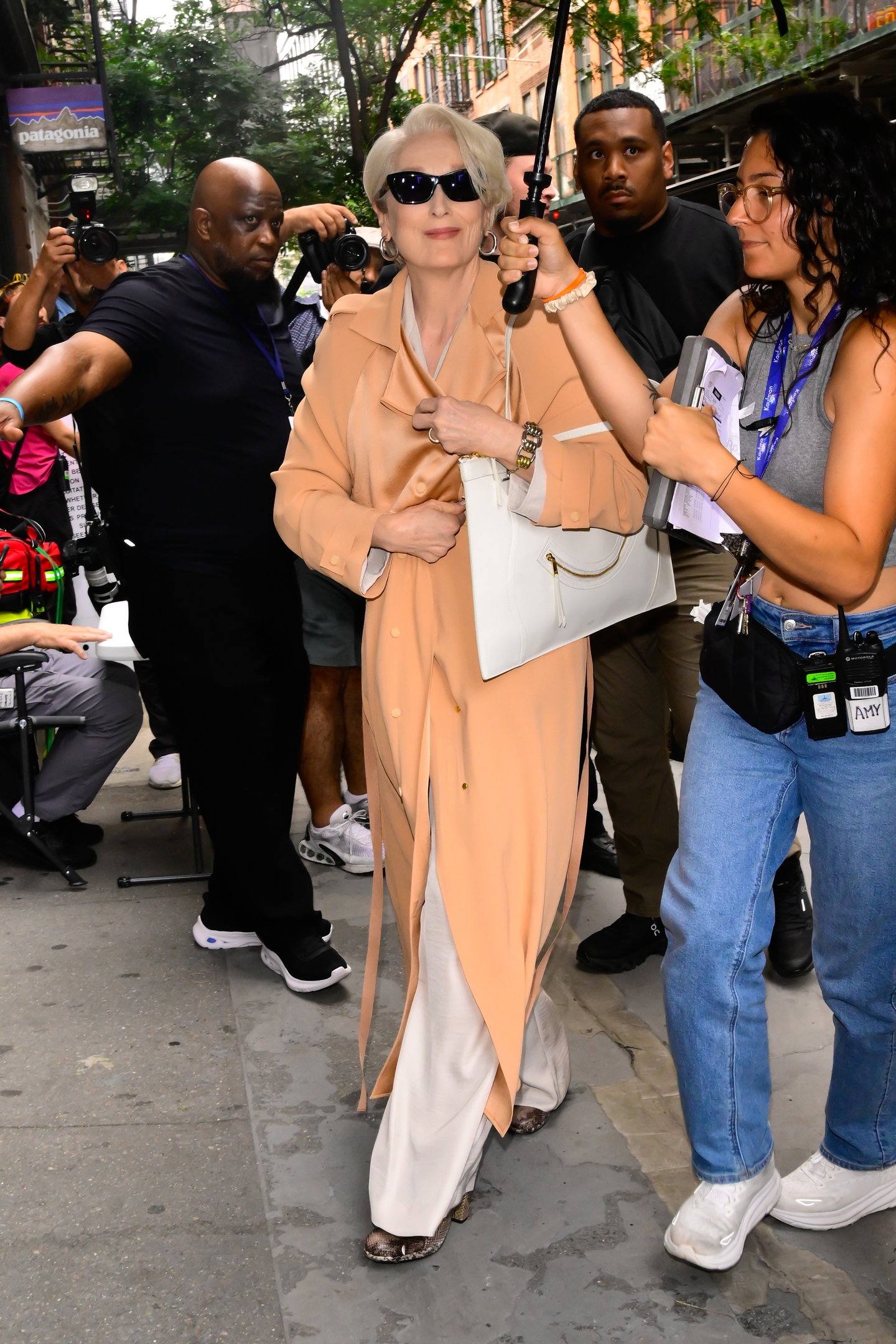 Uma mulher elegante de óculos escuros andando na rua com seguranças e fotógrafos ao redor.