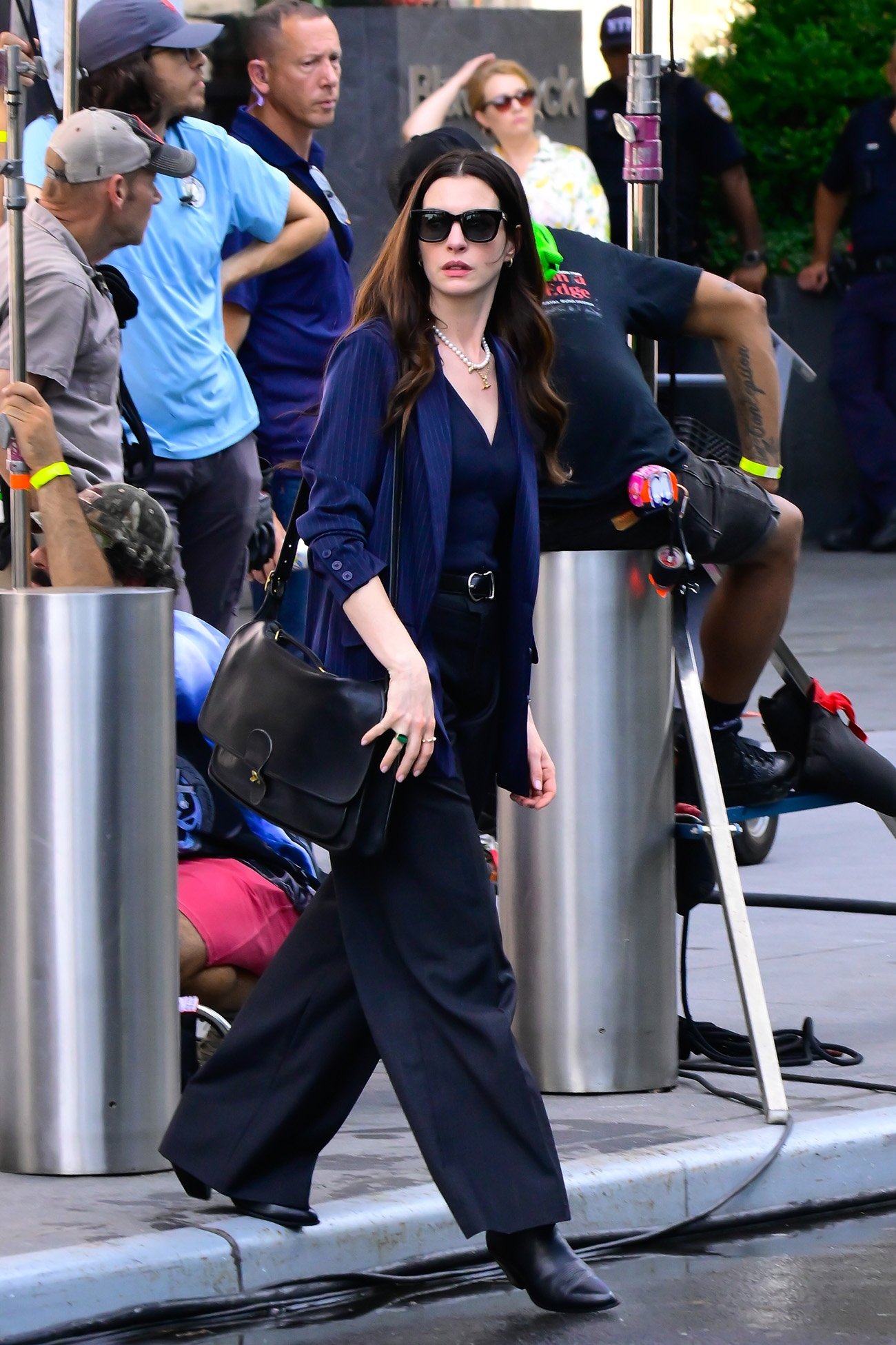 Pessoa em trajes elegantes caminha em um set de filmagem, cercada por equipe de produção. Sugerindo estilo O Diabo Veste Prada.