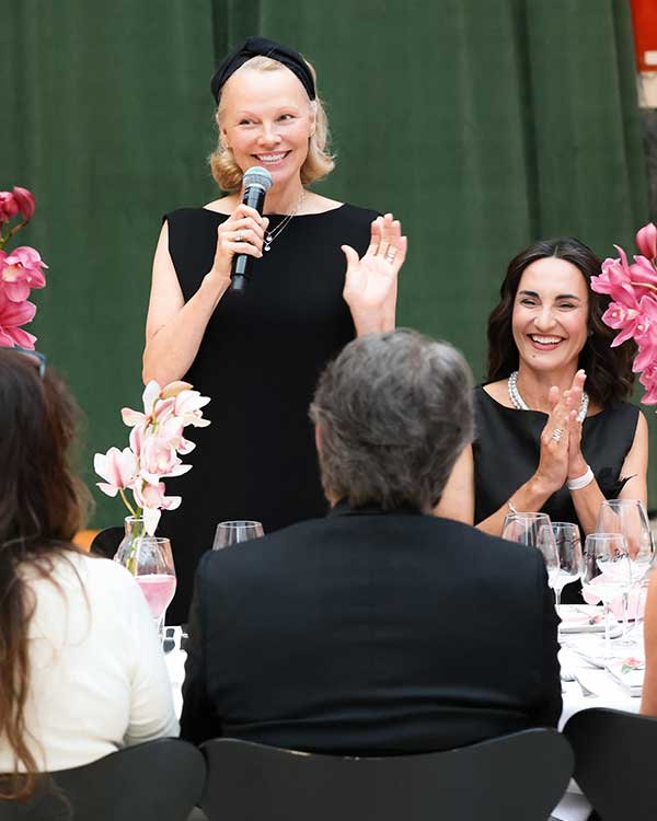 Pamela Anderson, de vestido preto ao centro, fala ao microfone em uma mesa cercada por convidados sorridentes.