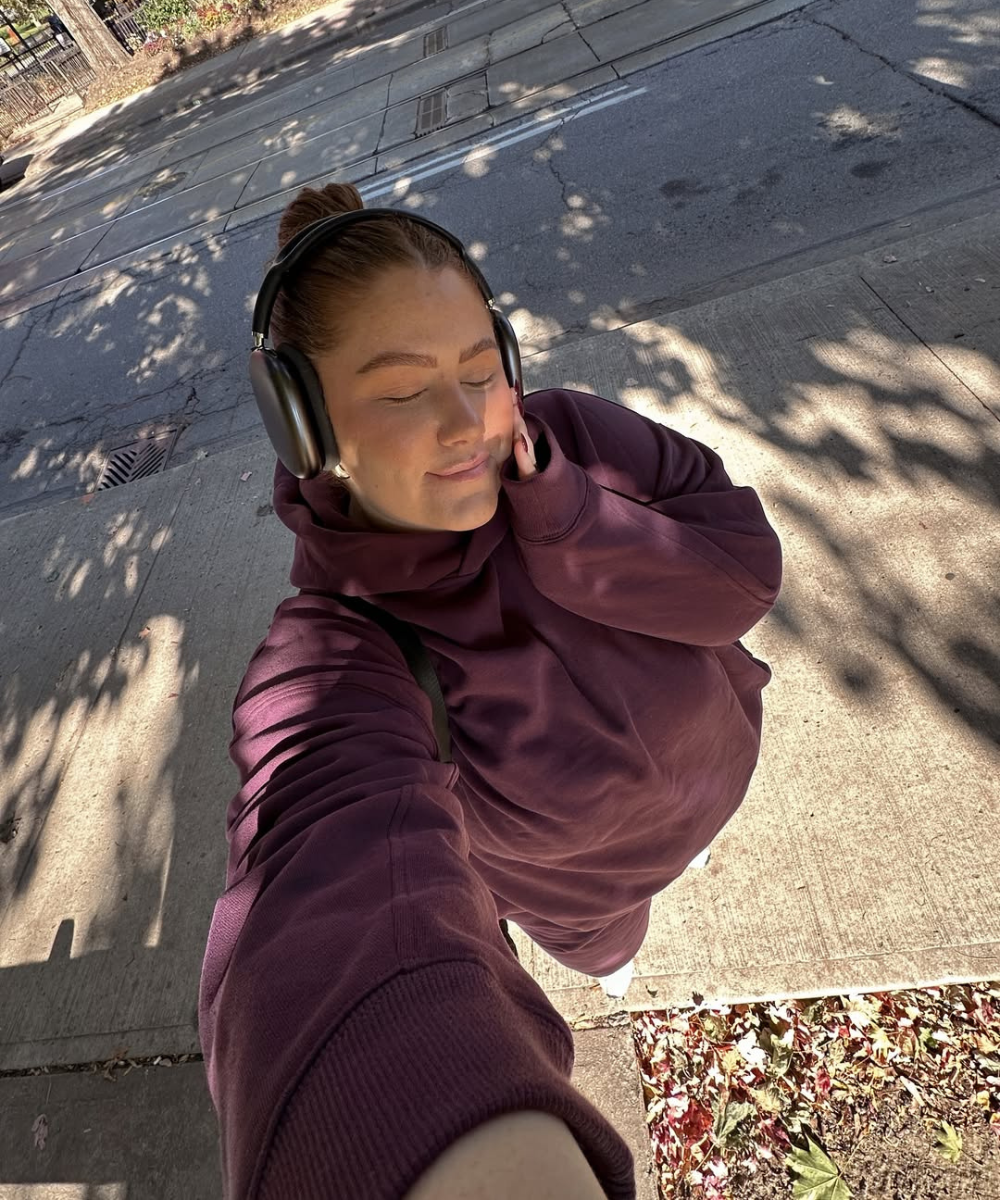 Mulher com fones e moletom roxo em rua arborizada, aproveitando o momento ao ar livre.