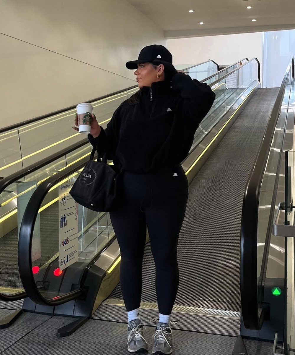 Pessoa de roupa esportiva preta, segurando café e usando boné, em uma escada rolante.