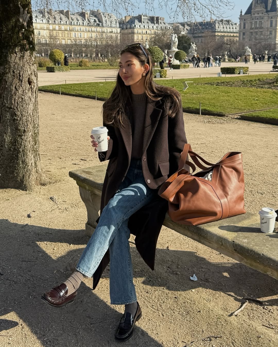 Mulher sentada em banco de parque, segurando café. Veste casaco e calça jeans, bolsa ao lado. Ao fundo, jardim e edifícios.