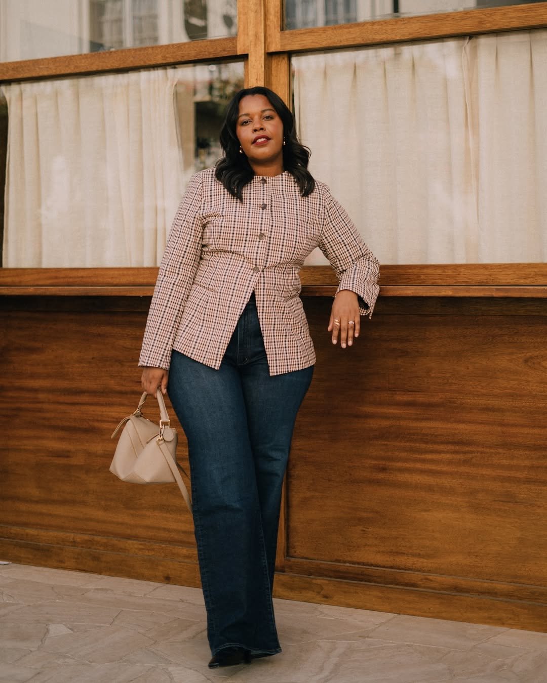 Mulher usando blazer xadrez e jeans escuro, demonstrando como usar jeans no trabalho. Ela segura bolsa clara.