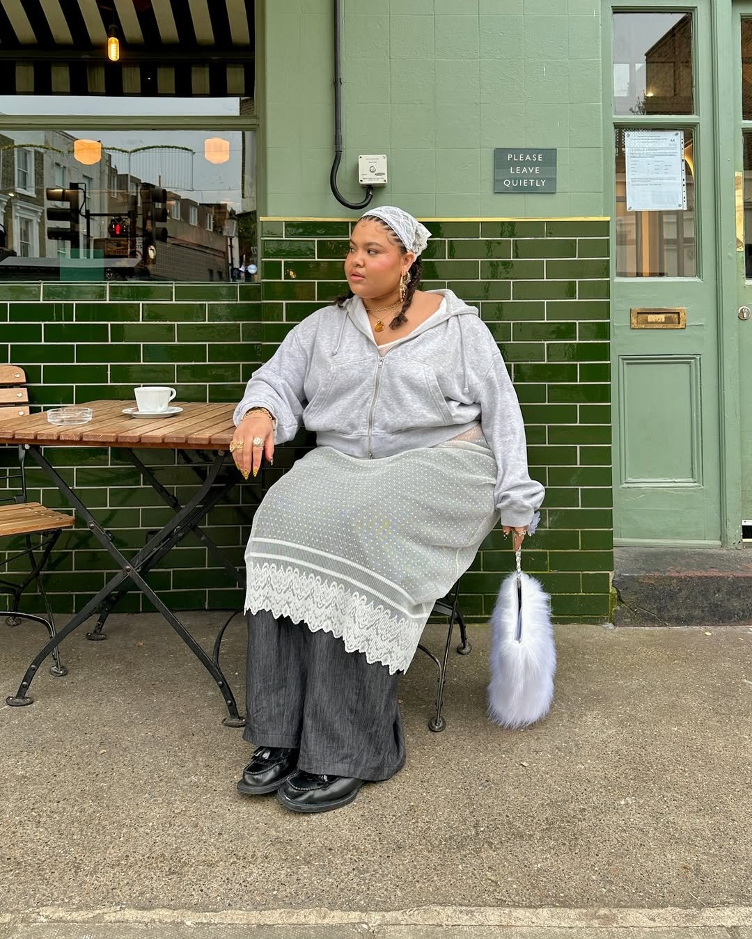 Pessoa sentada em uma mesa de café, usando um lenço de renda na cabeça, moletom cinza e saia sobre calça larga. Ela segura uma bolsa felpuda.